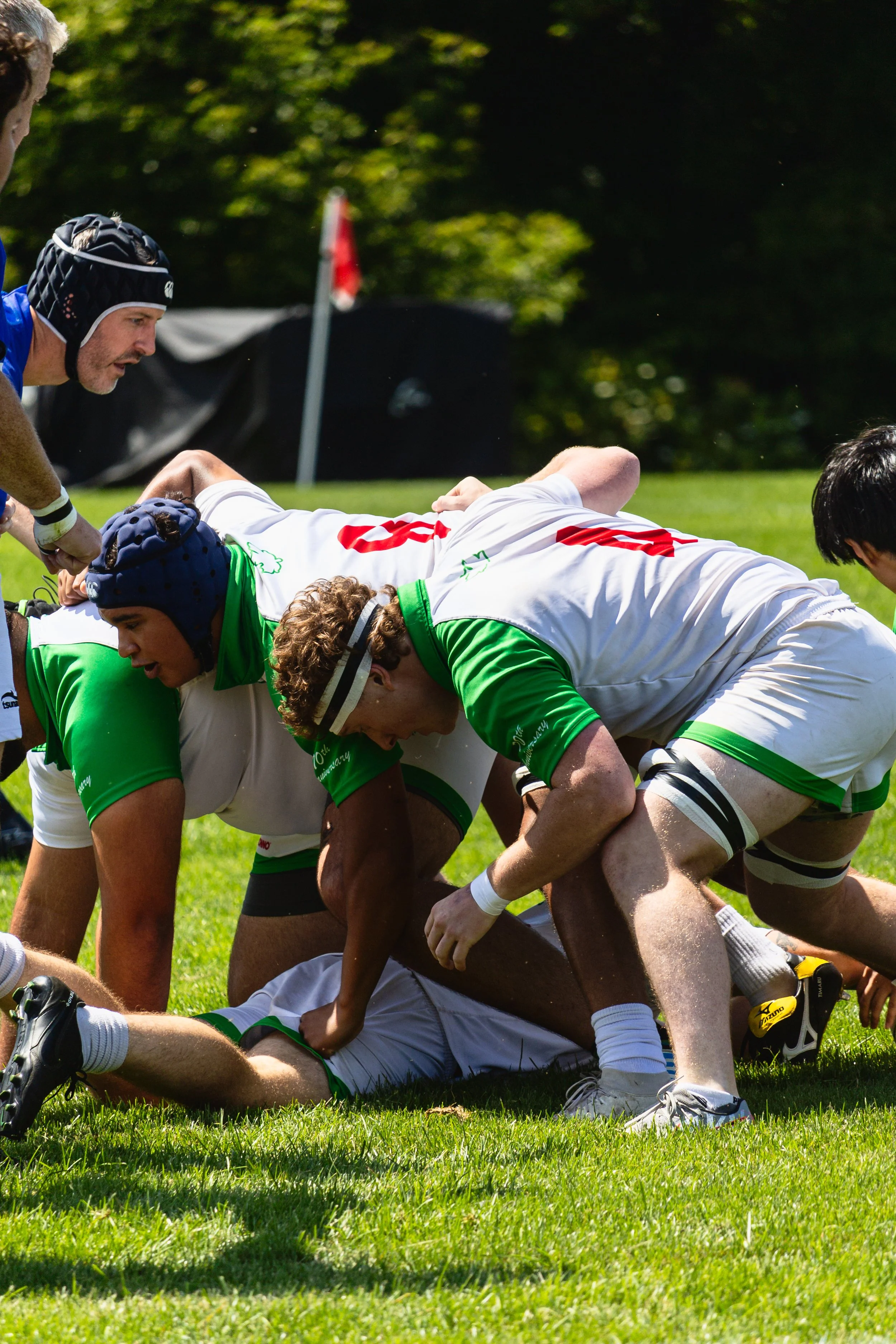 Rugby players in green and white uniforms forming a scrum on the field during a match, with one player on the ground beneath others, and a referee or coach observing nearby.