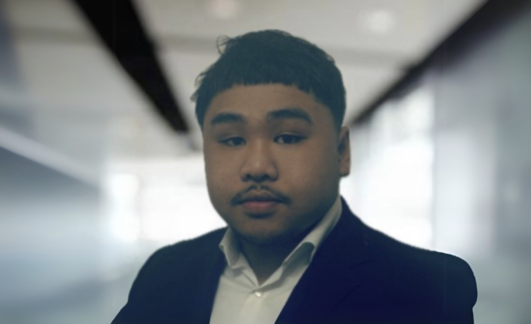 A young Asian man in a formal suit and white shirt looks directly at the camera with a neutral expression in an indoor setting with blurred background.