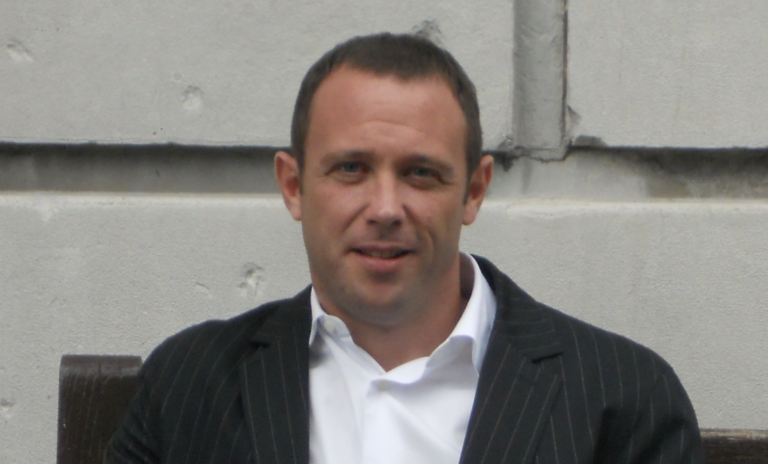 A man with short dark hair, wearing a black pinstripe suit and a white dress shirt, sitting outdoors against a concrete wall.