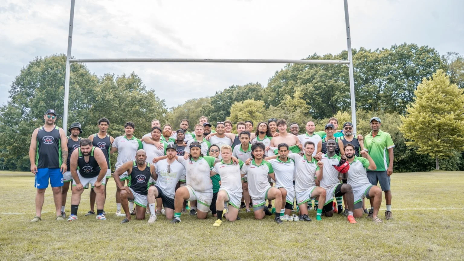 Group photo of rugby players and coaching staff on a field with rugby goalposts and trees in the background.
