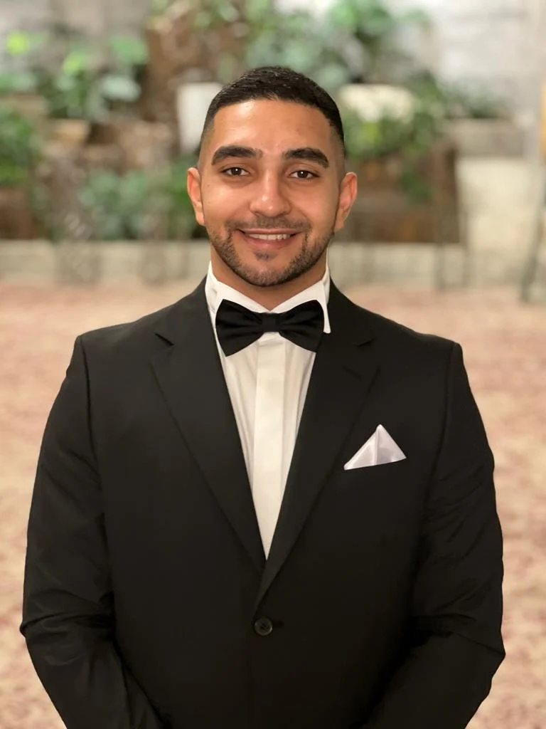 A young man in formal black tuxedo with a bow tie and white pocket square, smiling at the camera, standing indoors with blurred greenery in the background.