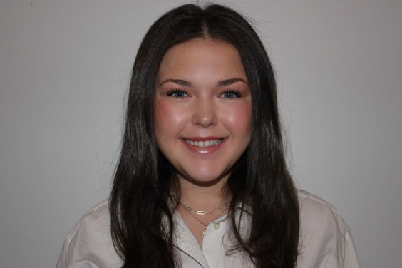 Young woman with long dark hair smiling at camera, wearing a white shirt and necklace, against a gray background.