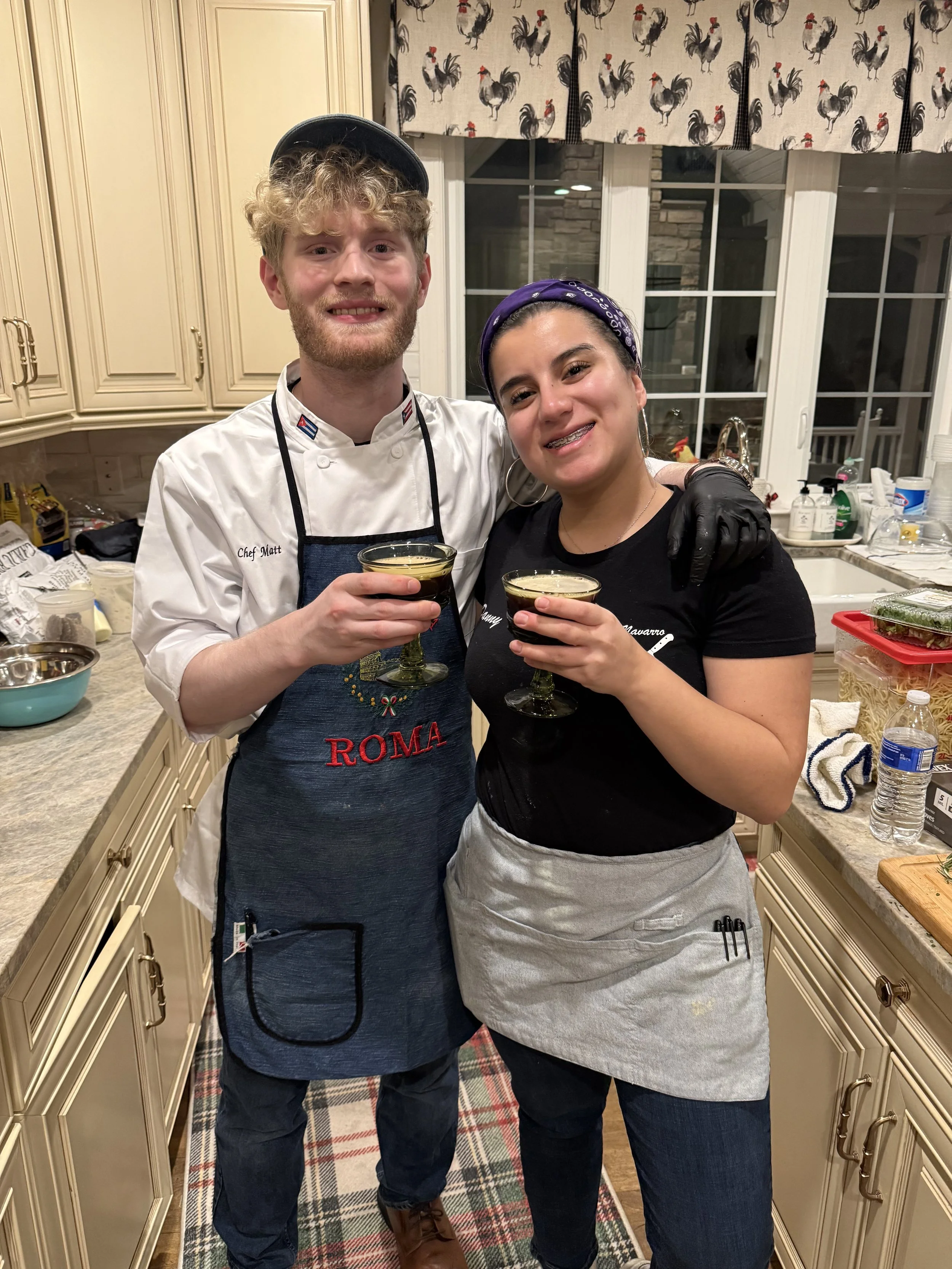 Two people in a kitchen, a man and a woman, holding glasses of dark-colored beverages. The man is wearing a white chef's coat and a black apron that says 'ROMA', and the woman is wearing a black shirt and a gray apron. They are smiling and standing close together.