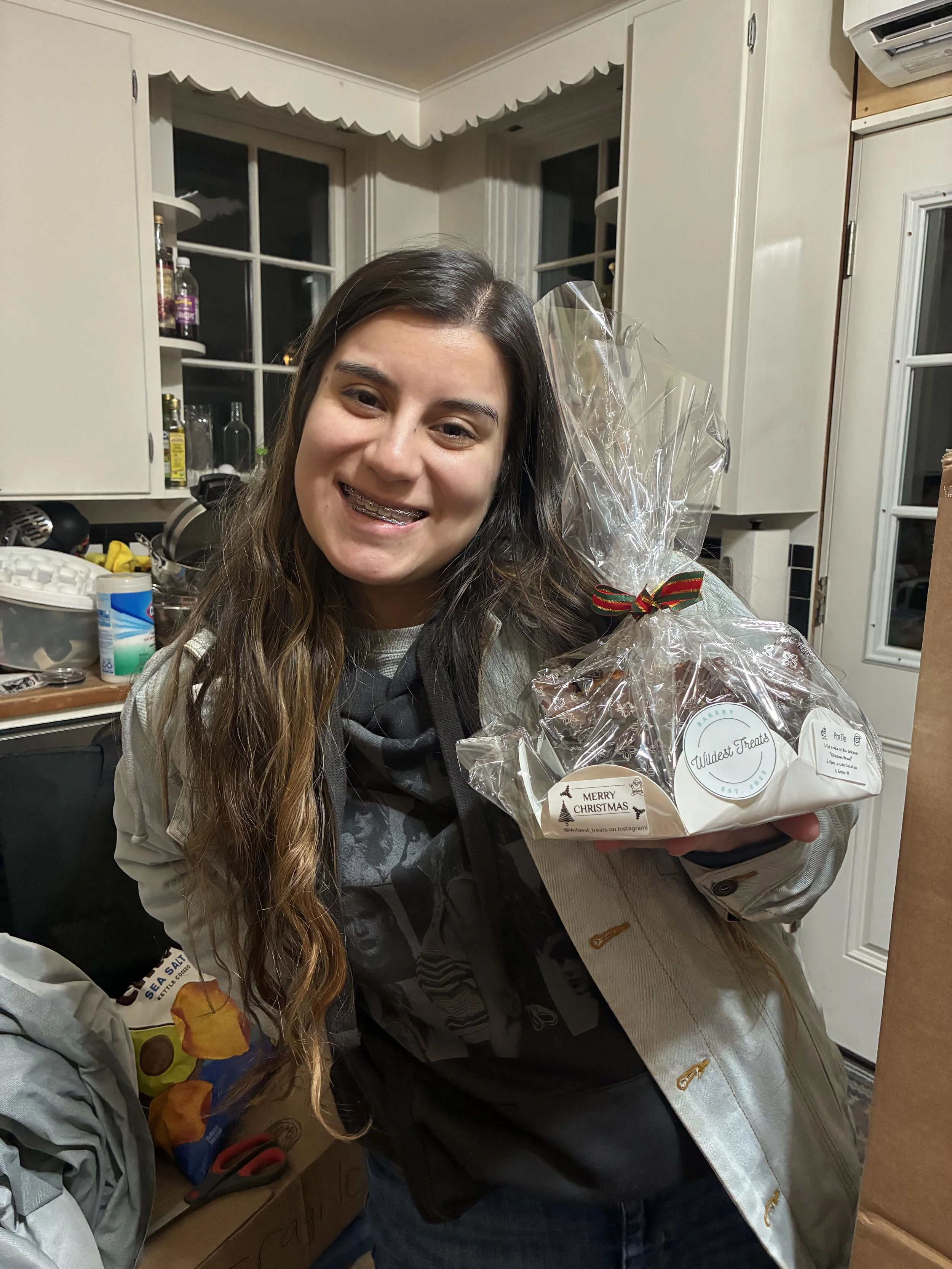 A young woman with long brown hair smiling while holding a clear plastic-wrapped gift basket with Christmas tags inside a kitchen.