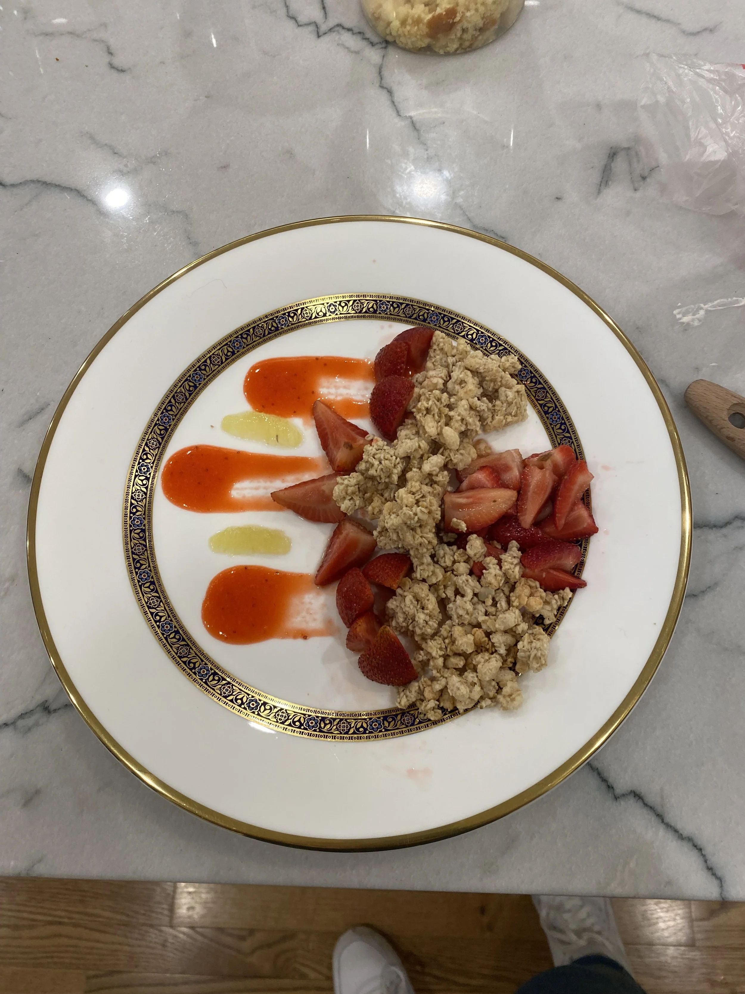A decorative white plate with gold trim holding fresh sliced strawberries, strawberry slices with granola, and dollops of red and yellow sauces on a marble tabletop.