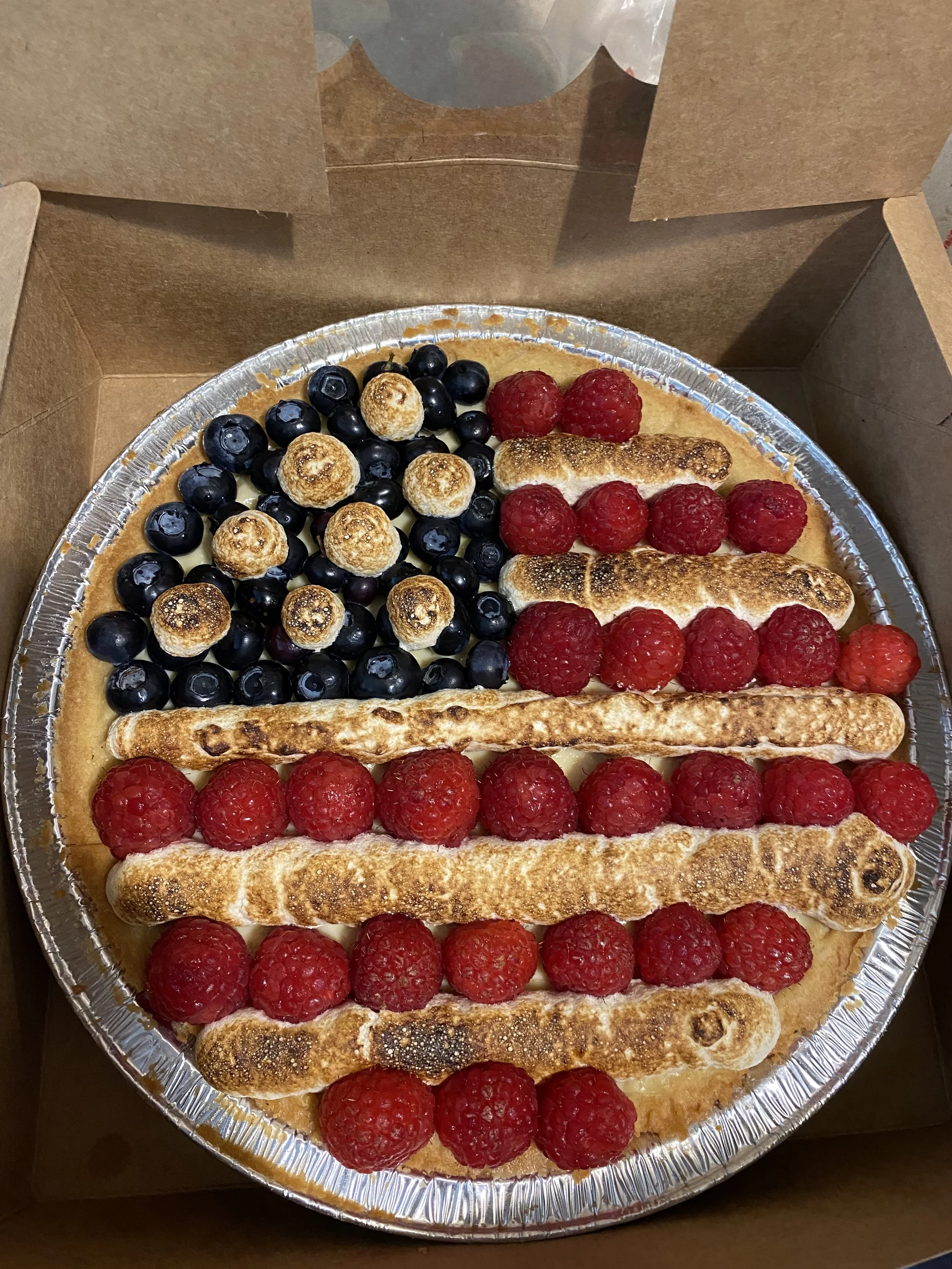 A fruit tart with blueberries and toasted meringue on top of a crust, decorated with fresh raspberries and ladyfingers, in a cardboard box.