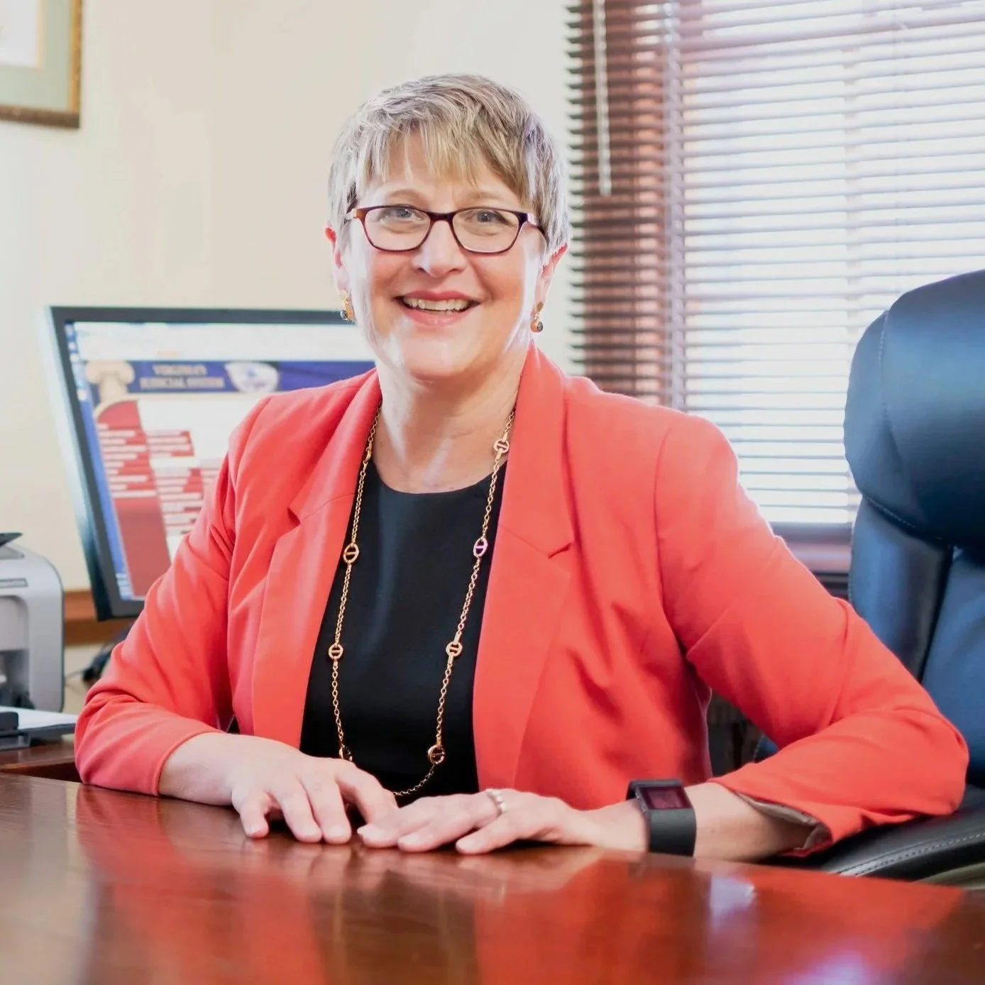 A smiling woman wearing glasses, a black top, a coral blazer, and gold jewelry sitting at a desk with a computer and window blinds in the background.