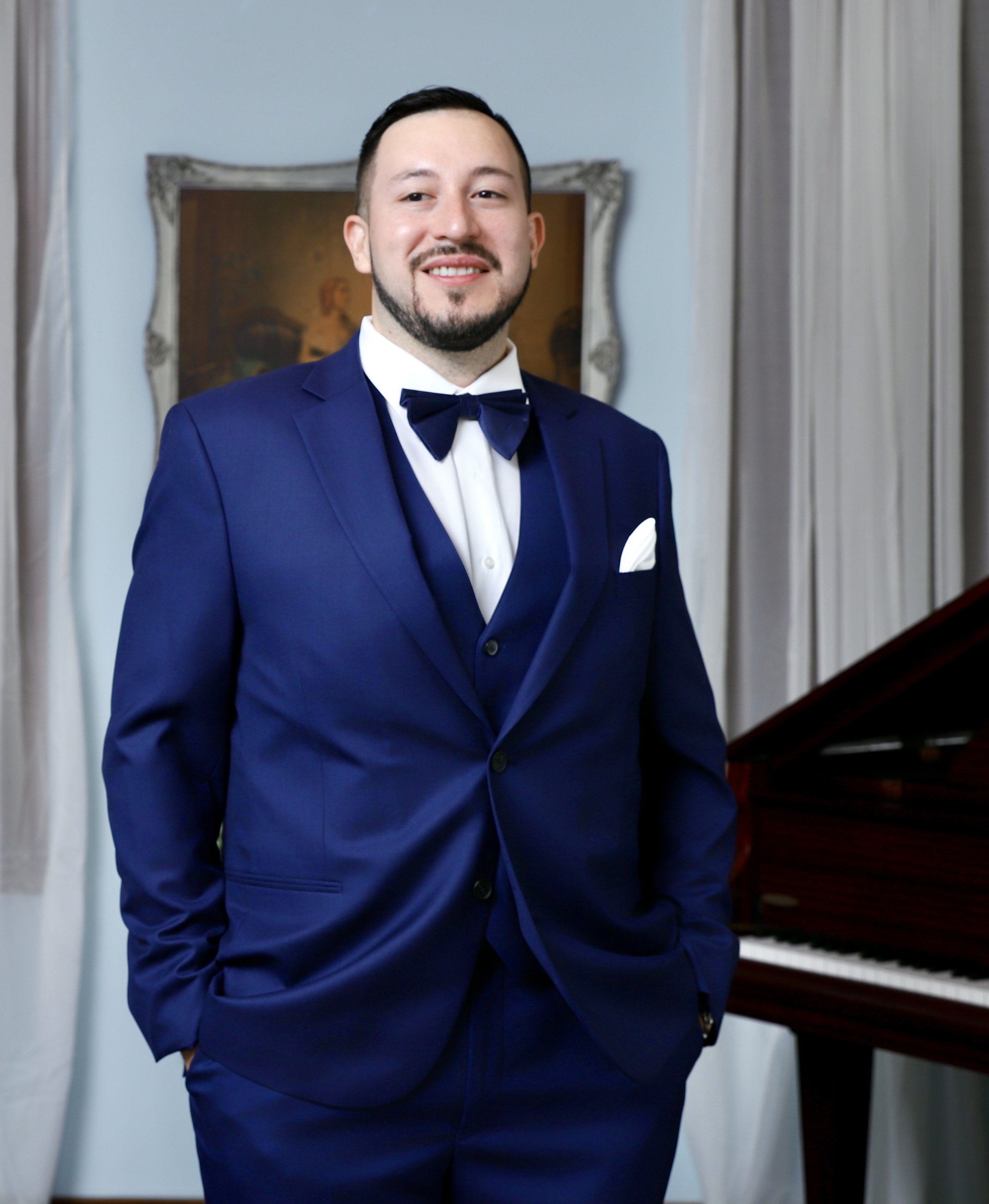 A man dressed in a blue tuxedo with a black bow tie and white shirt, standing indoors in front of a mirror and a piano, smiling at the camera.