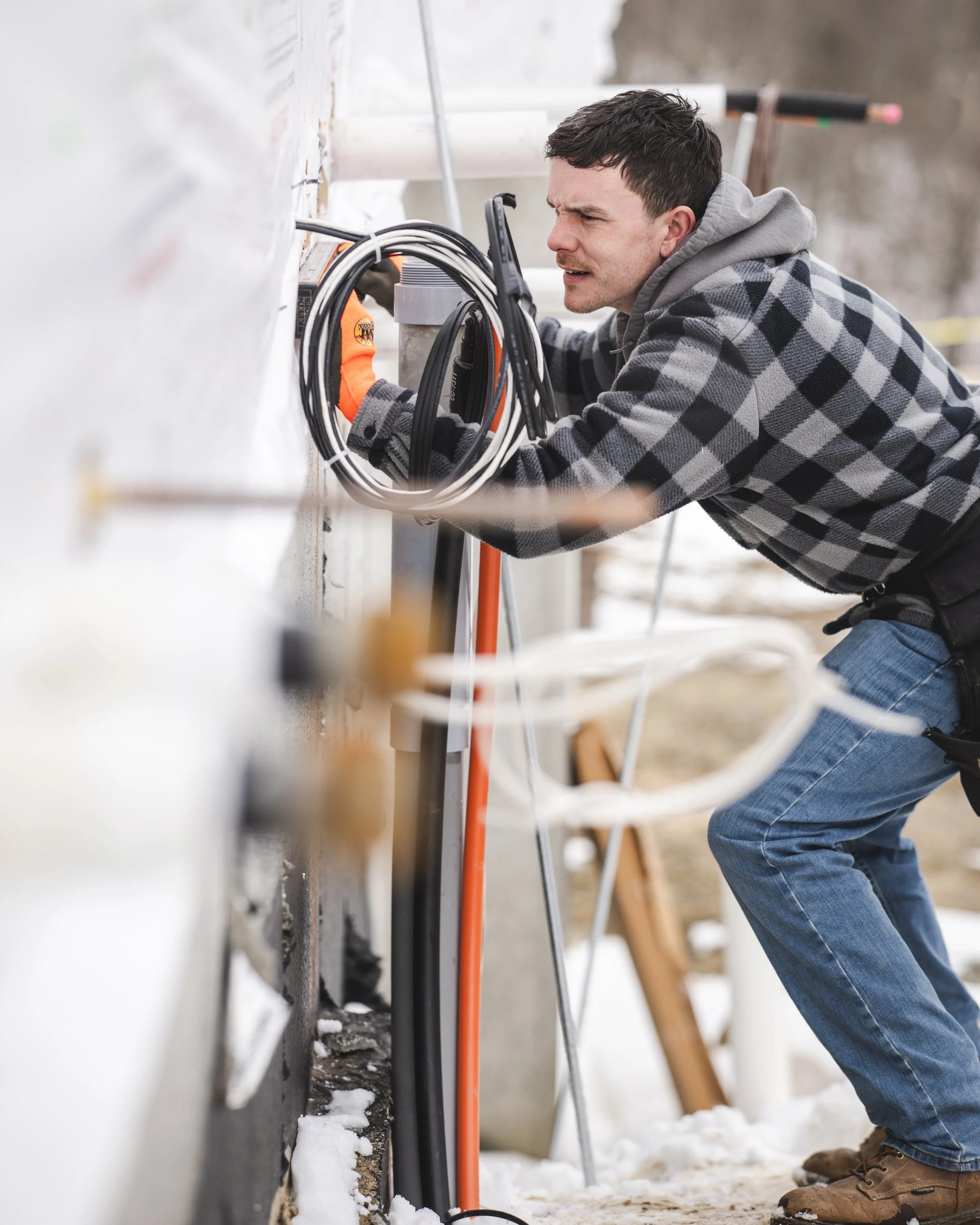 A man in a checkered jacket is working on electrical wiring outside in winter, crouching near a wall with snow on the ground.