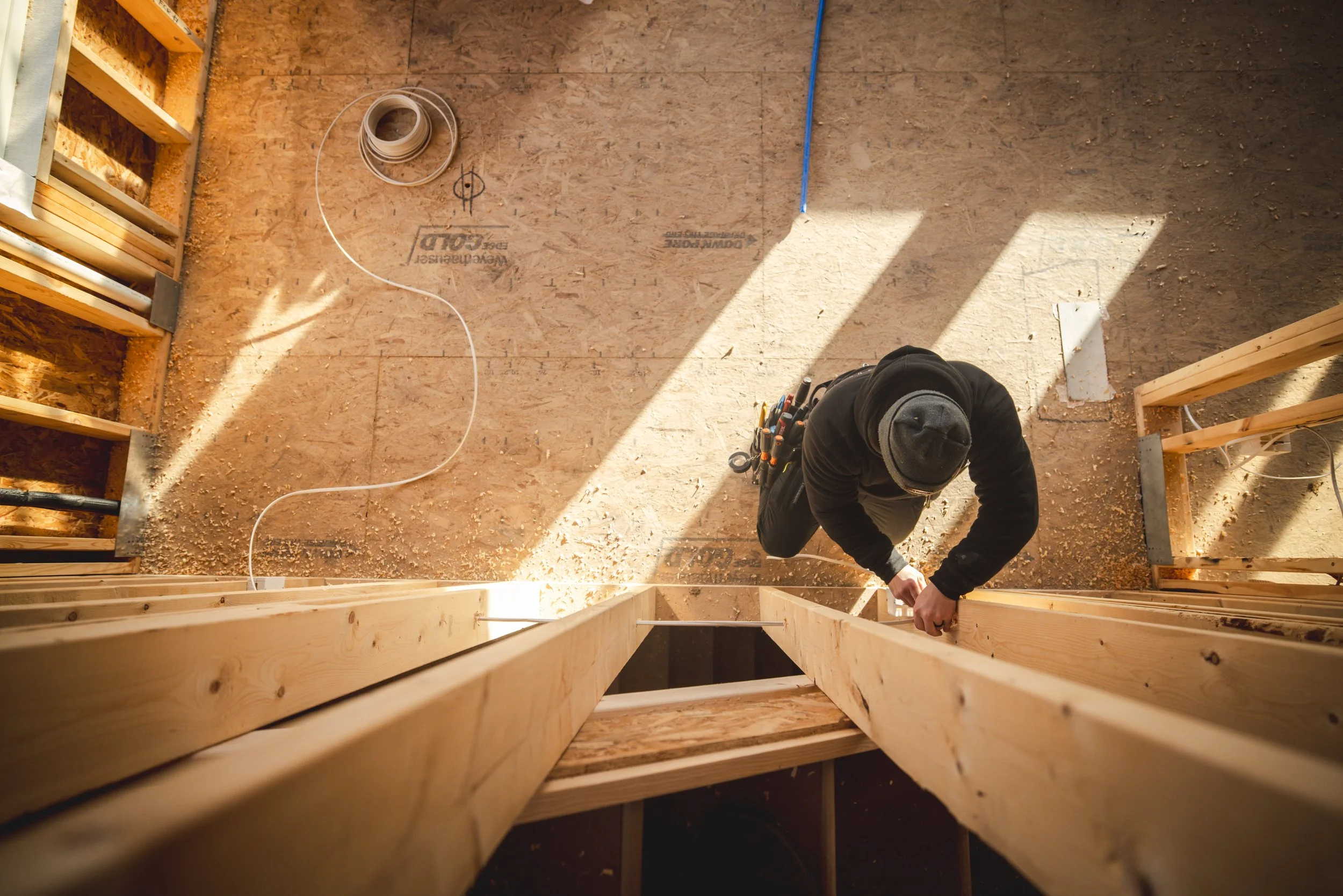 A construction worker wearing a black hoodie, gray beanie, and tool belt is working on wooden framing inside a building under construction, seen from above with sunlight shining through.