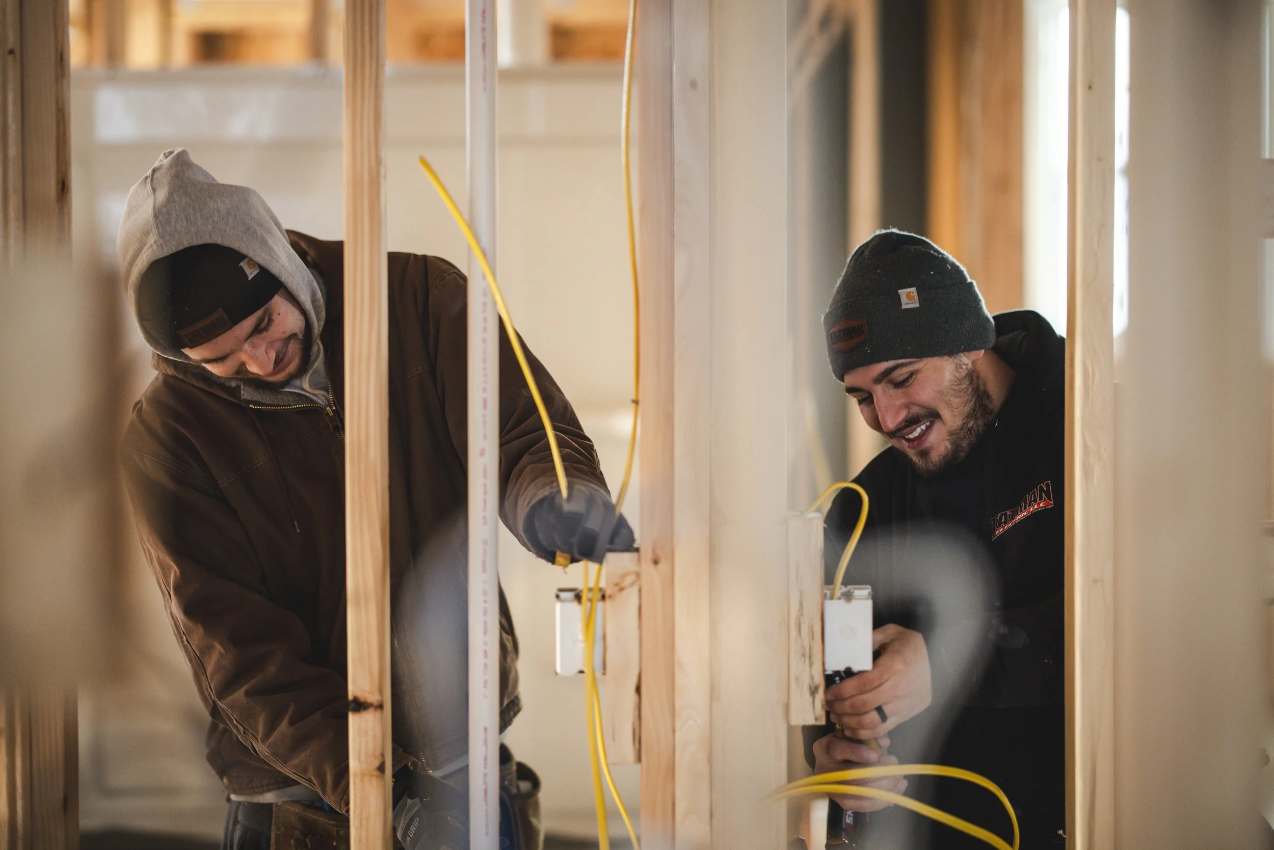 Two men working with electrical wiring inside framed wooden wall studs in a construction site, smiling and wearing warm clothing and beanies.