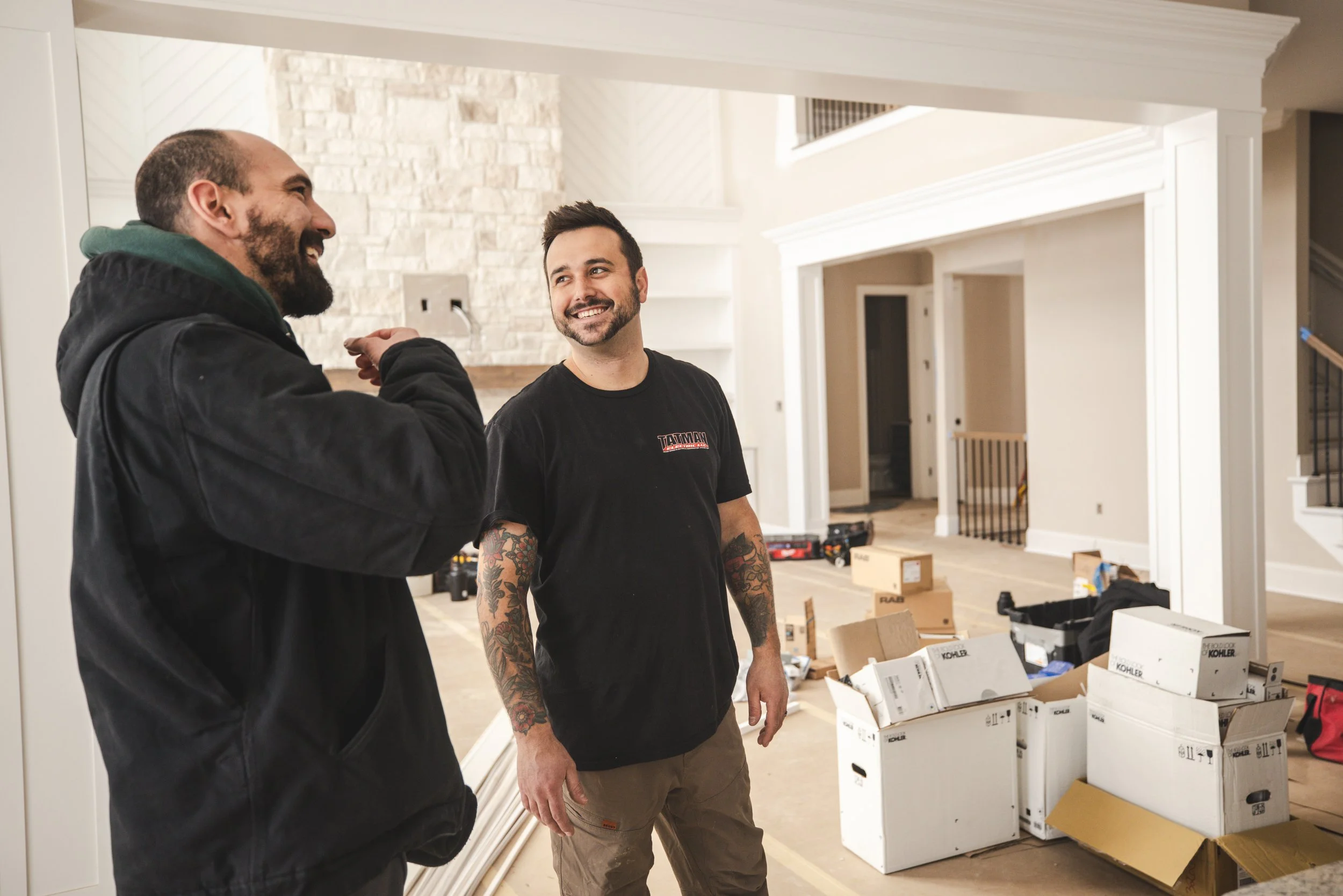 Two men are smiling and talking inside a house undergoing renovation. One is wearing a black hoodie and the other a black T-shirt with tattoos on his arms. There are open boxes and renovation tools around them, with a partially finished interior in the background.