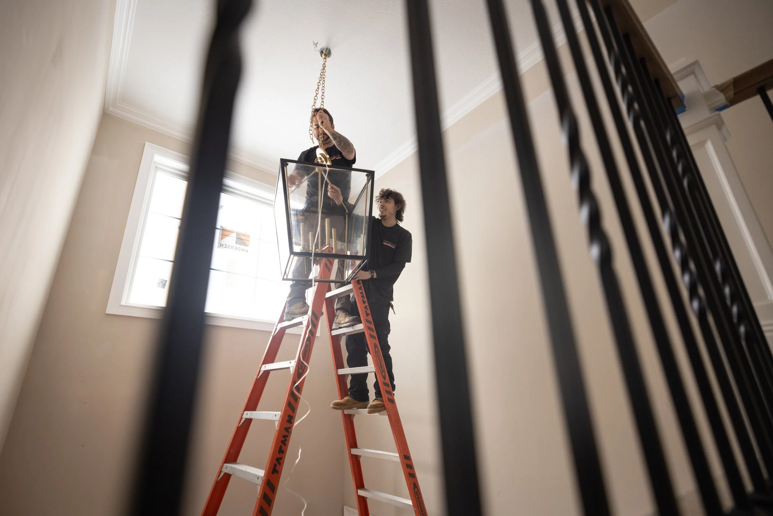Two men install a chandelier inside a home, standing on a red ladder in front of a large window, viewed through black staircase bars.