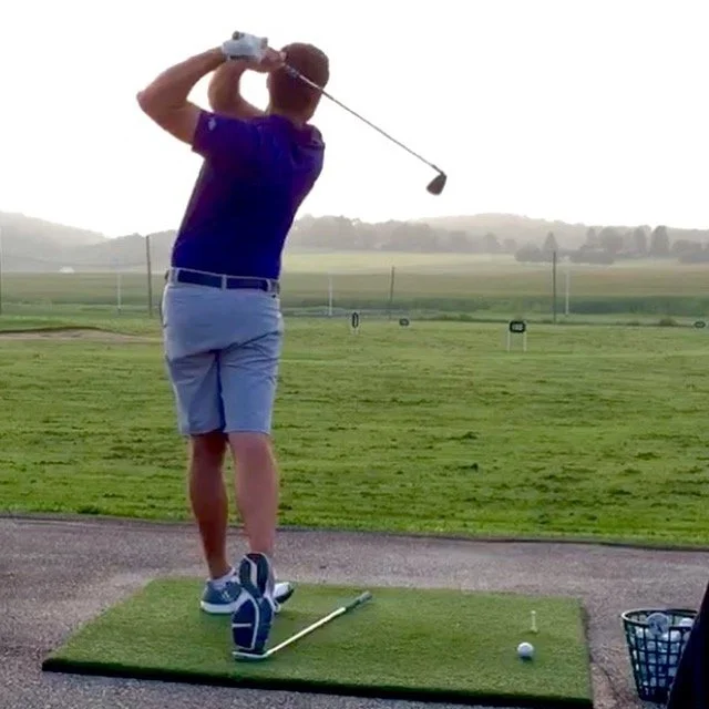 A person in a blue shirt and gray shorts practicing golf swings on a field at a golf driving range, with a golf club in hand and a golf ball on a tee.