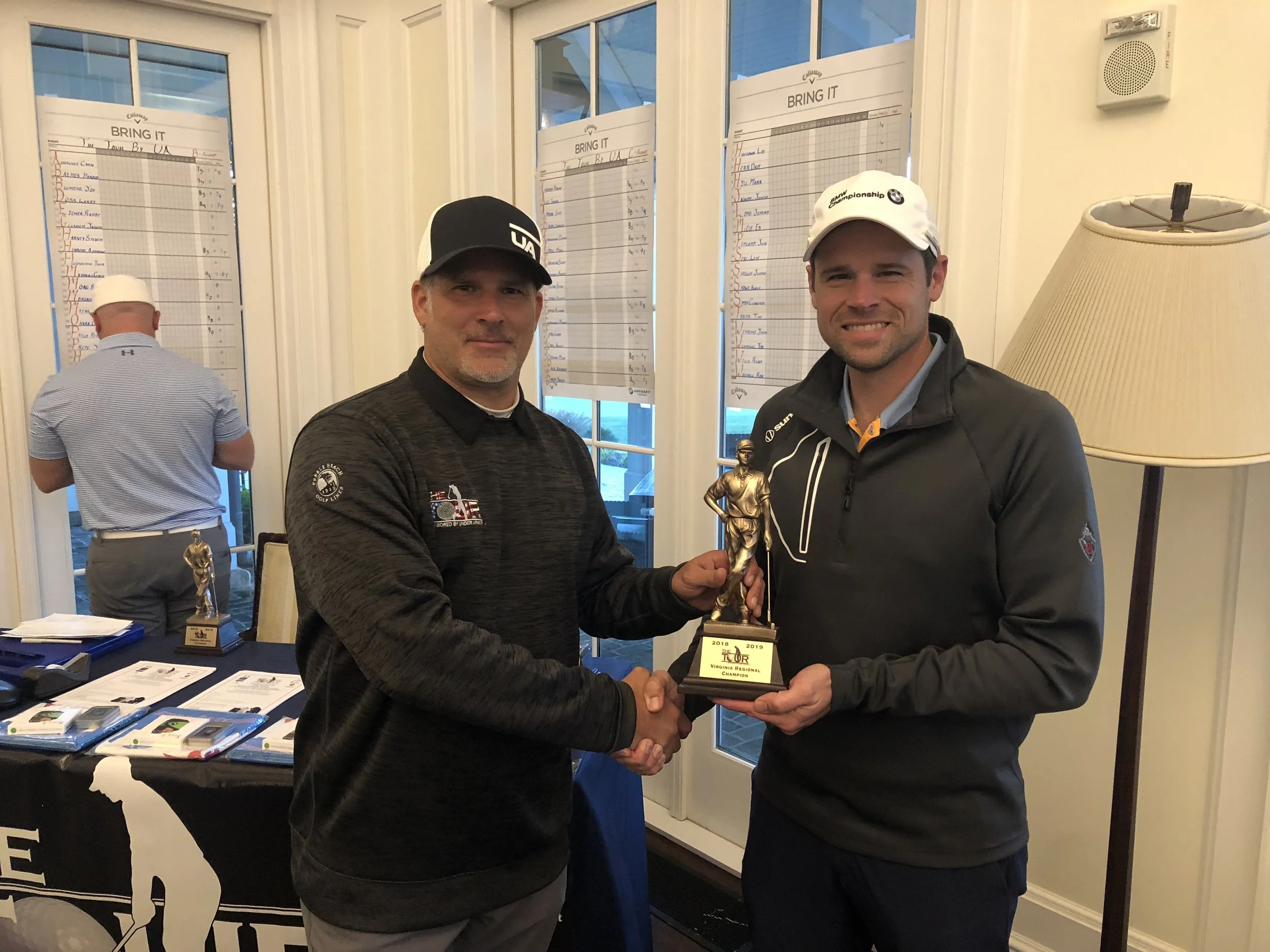 Two men in golf attire shaking hands, one is receiving a trophy, in a room with a large window and a table with papers and a smaller trophy on it.