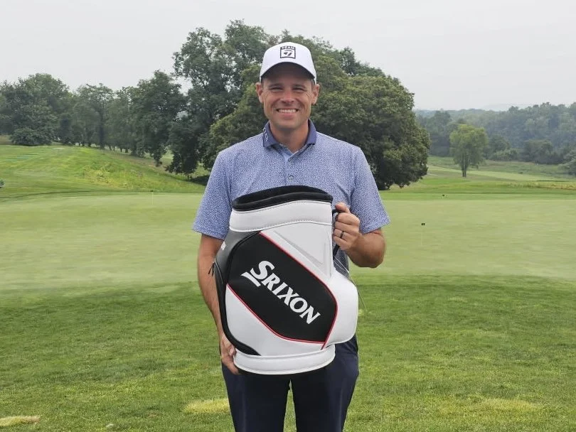 A man standing on a golf course holding a Srixon golf bag, wearing a white cap and a blue button-up shirt, with trees and greenery in the background.
