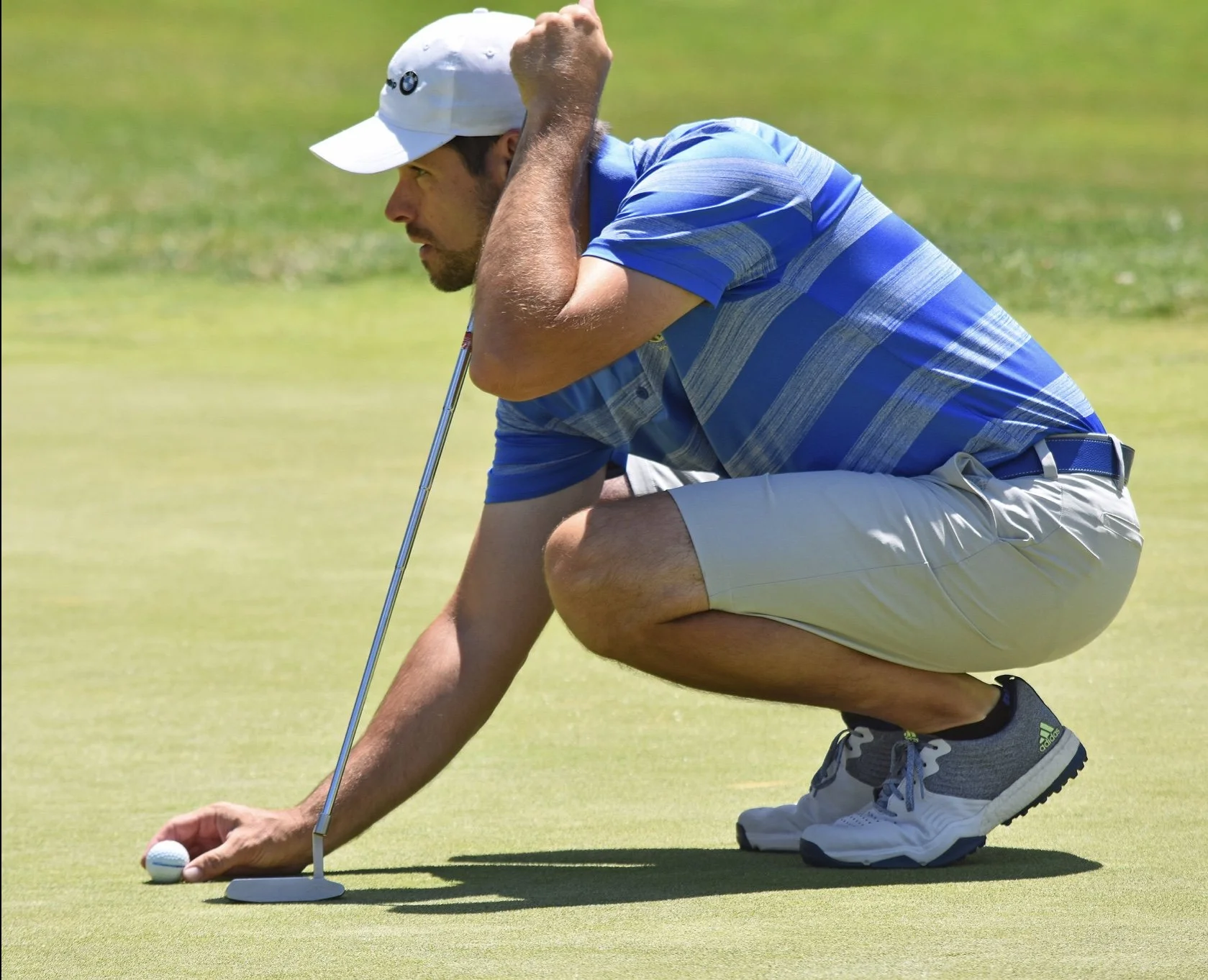 A man crouching on a golf course, lining up a putt with a golf club and ball.