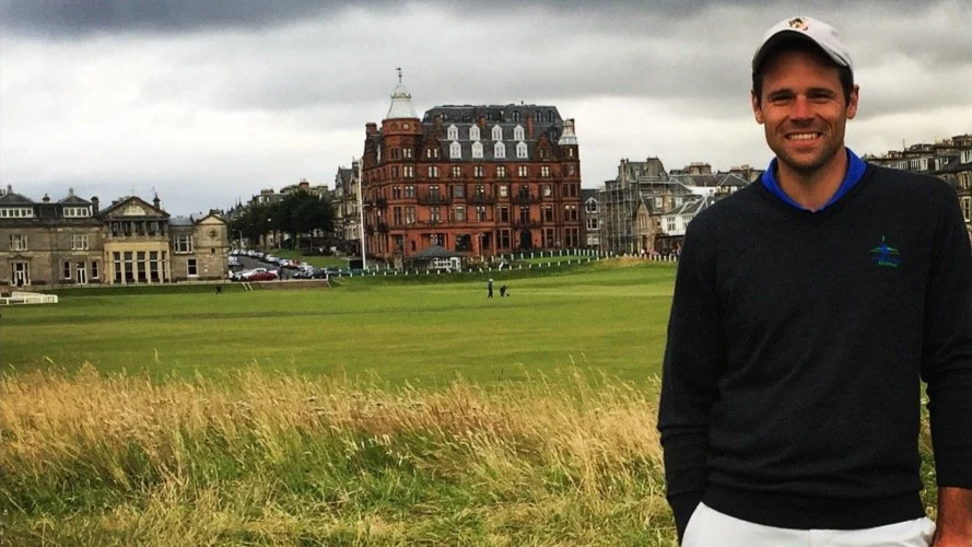 A man smiling, wearing a gray hat and black sweater, standing on a grassy area with historic buildings in the background.