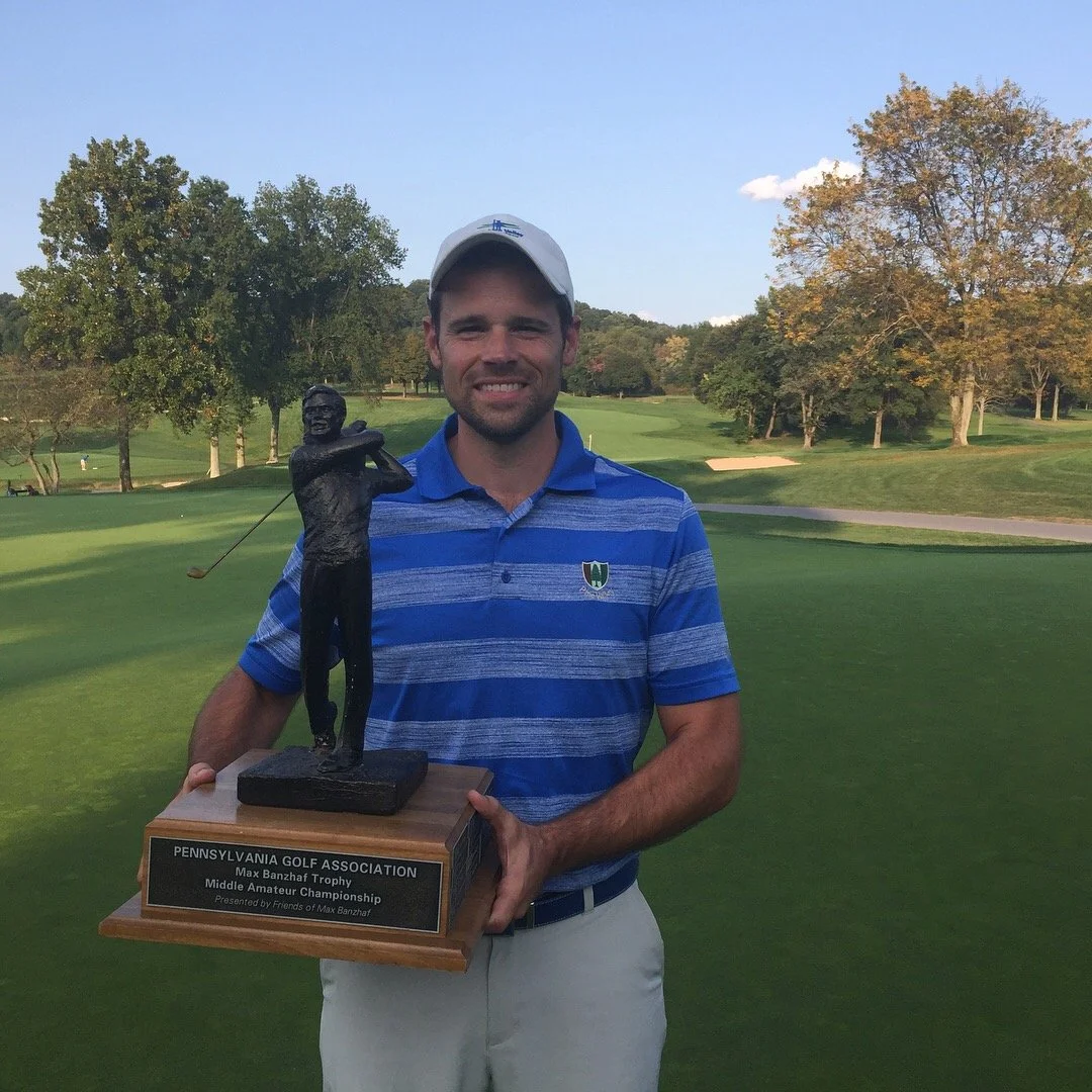 A man in a blue striped polo shirt and white cap holding a trophy on a golf course with trees and a blue sky in the background.