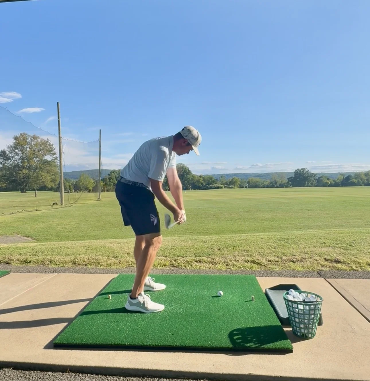A man practicing golf swing on a golf driving range, standing on a green mat, with a basket of golf balls nearby, under a clear blue sky.