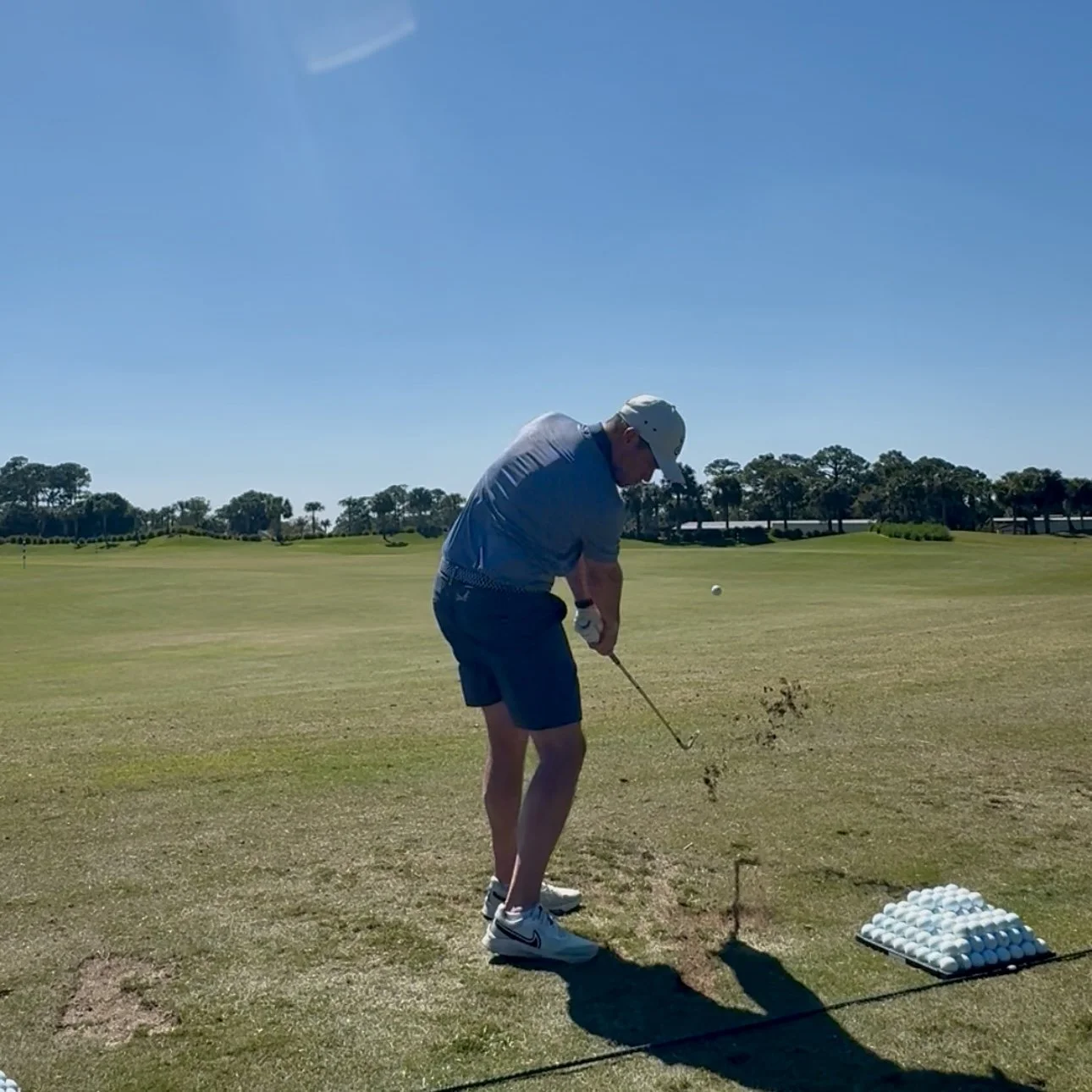 A man practicing golf on a sunny golf course, gearing up for a shot with a golf club, wearing a white hat, gray shirt, navy shorts, and white shoes, with a cluster of golf balls on a tee nearby.