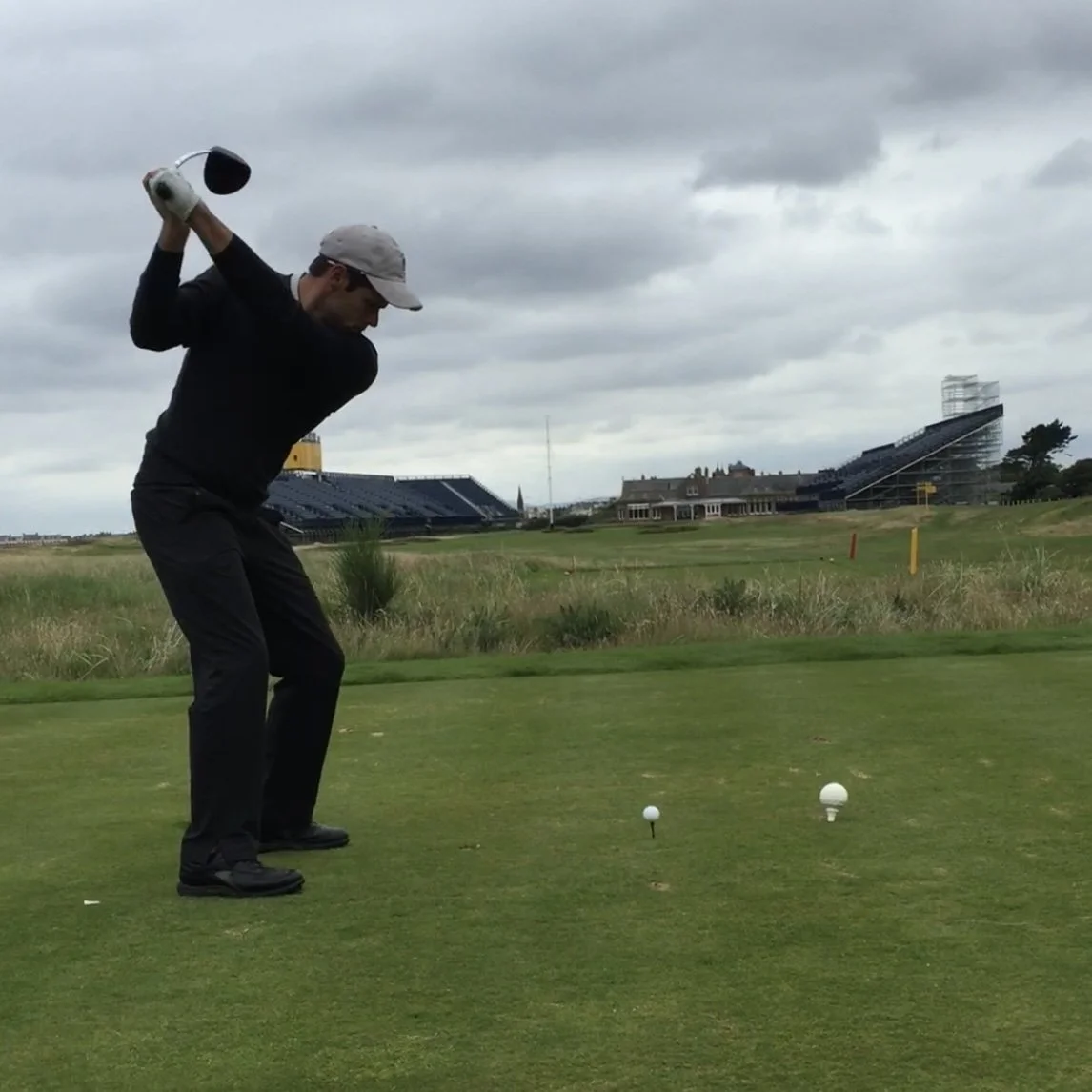 A man playing golf on a cloudy day, preparing to hit the ball from the tee box with golf clubs behind him and a golf course and grandstands in the background.