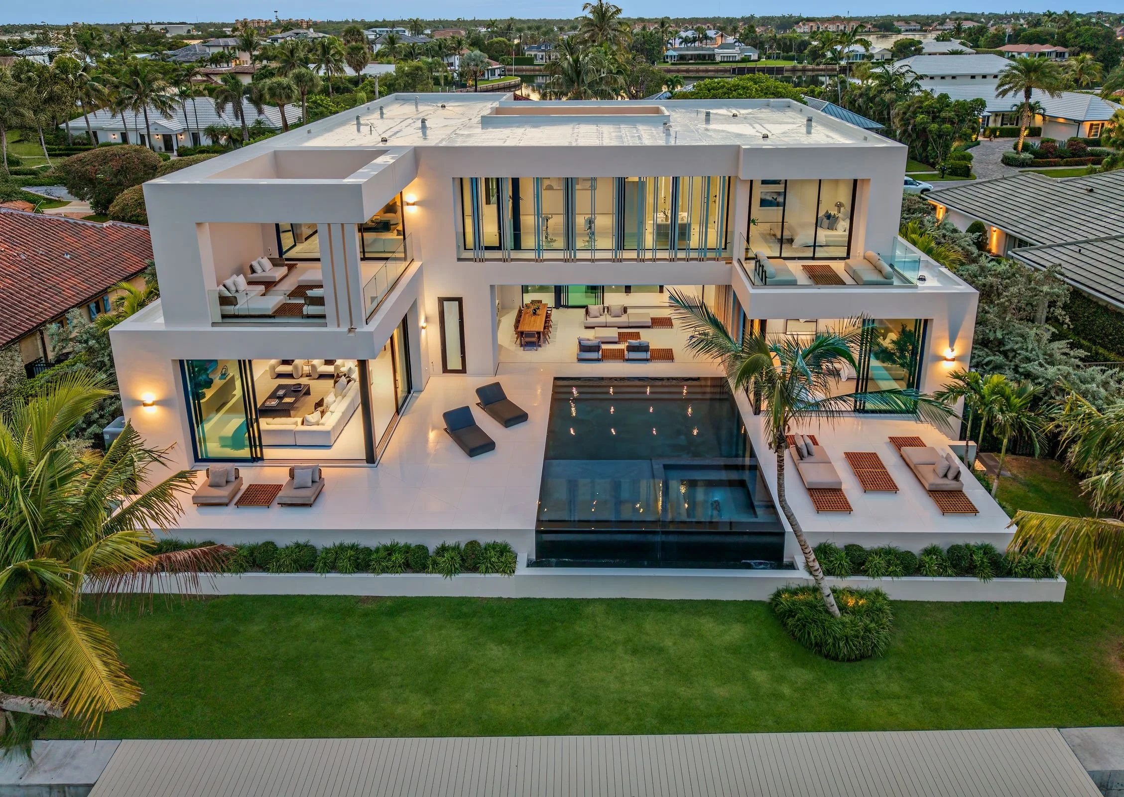 A modern, multi-story house with large glass windows, outdoor patio areas, a swimming pool with a black waterfall feature, lounge chairs, palm trees, well-manicured lawn, and surrounded by neighboring houses and greenery.