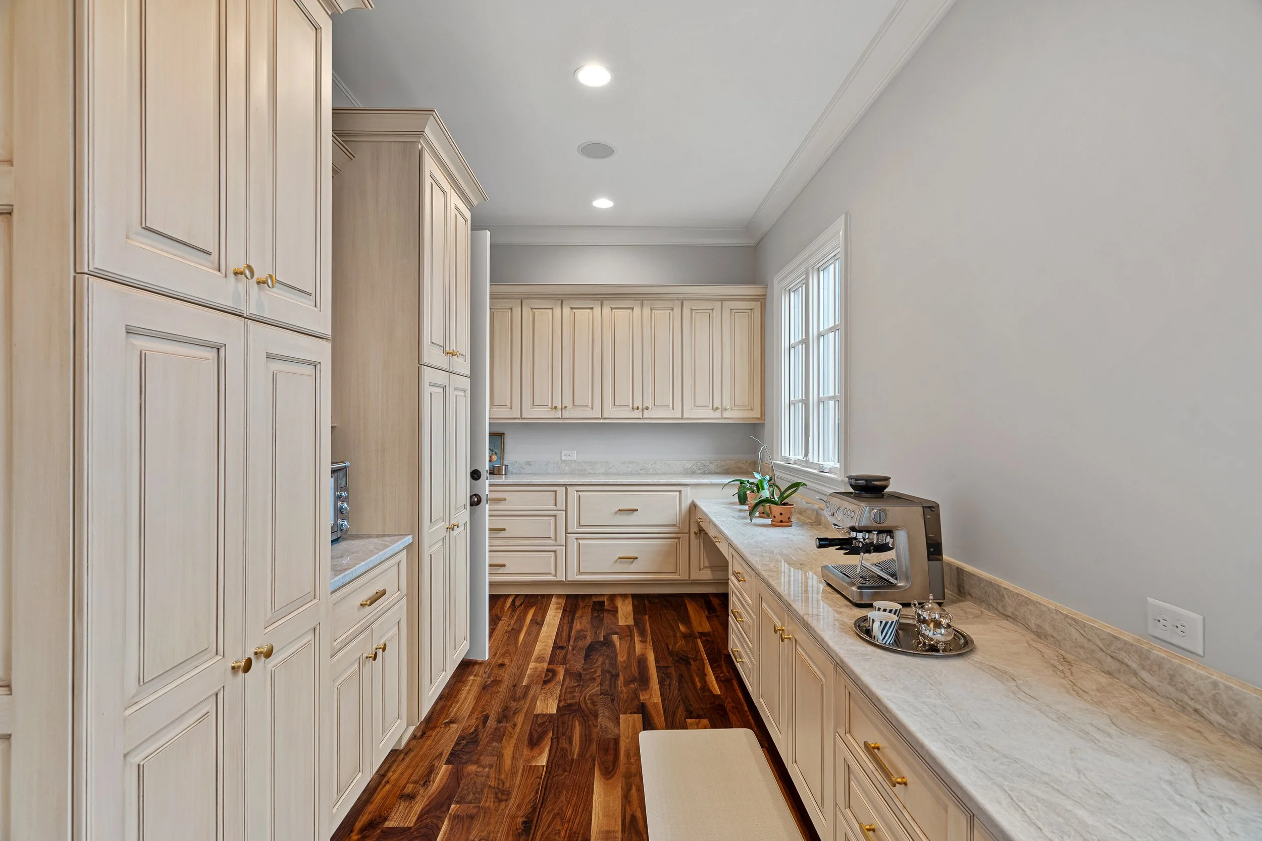 A kitchen with white cabinetry, marble countertops, and hardwood flooring. There are two large windows on the right, a coffee machine on the counter, and a small potted plant.