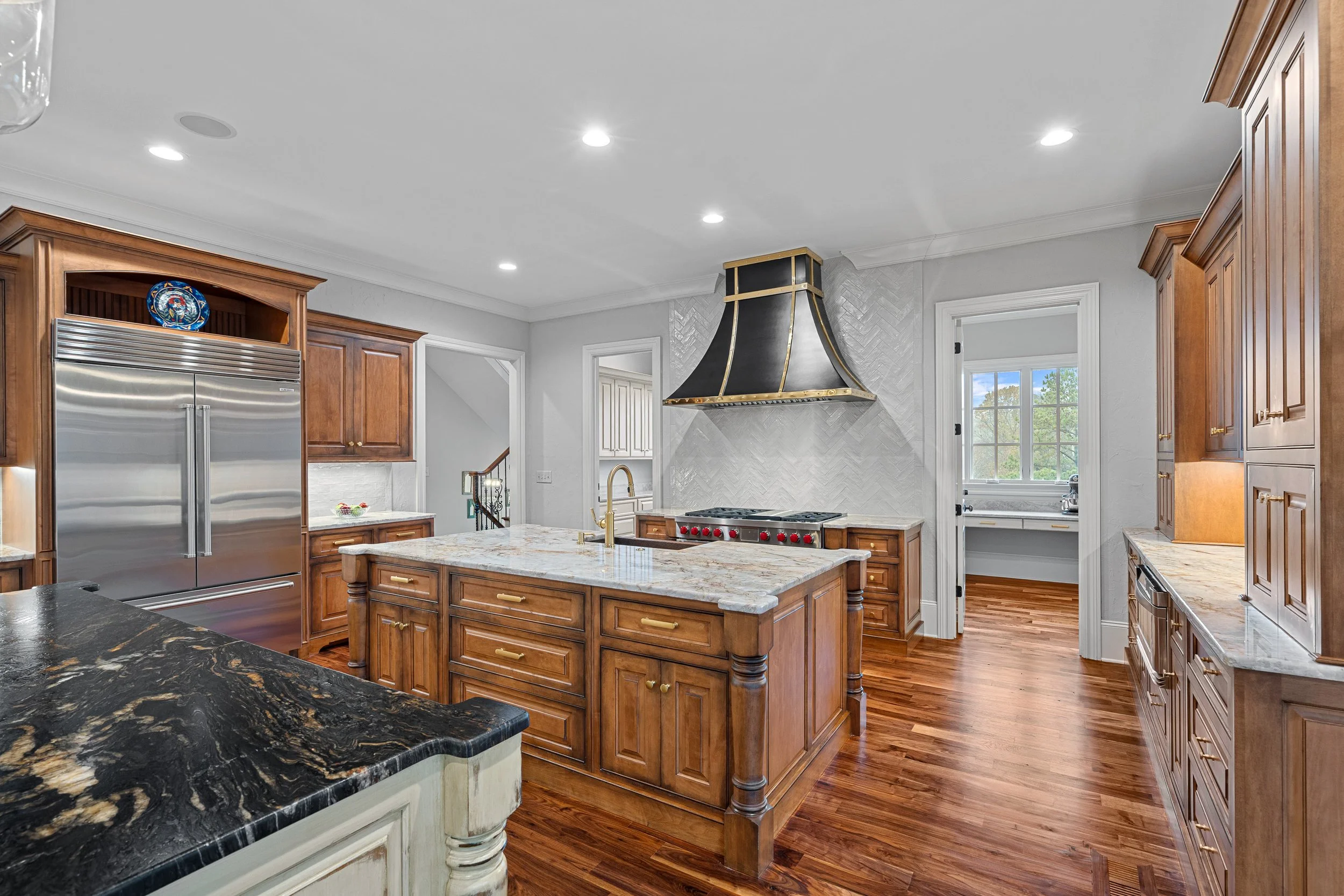 Spacious kitchen with wooden cabinetry, marble countertops, stainless steel refrigerator, and a large kitchen island with a gas stove and an ornate range hood. There is a small room with a window and a sink visible in the background.