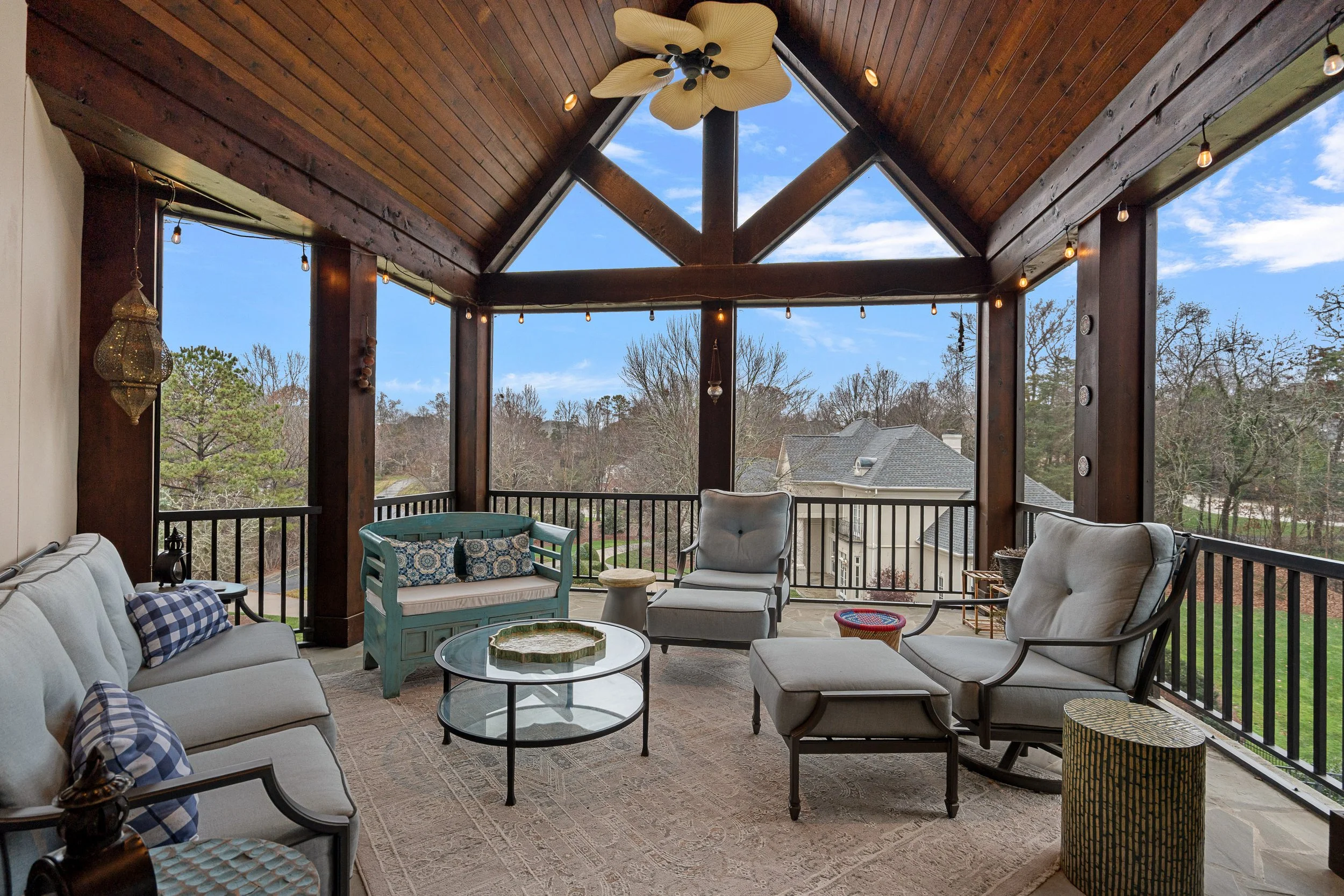 Covered porch with wooden ceiling, ceiling fan, string lights, and outdoor furniture including sofas, chairs, tables, and decorative cushions, overlooking a suburban neighborhood with trees and houses.
