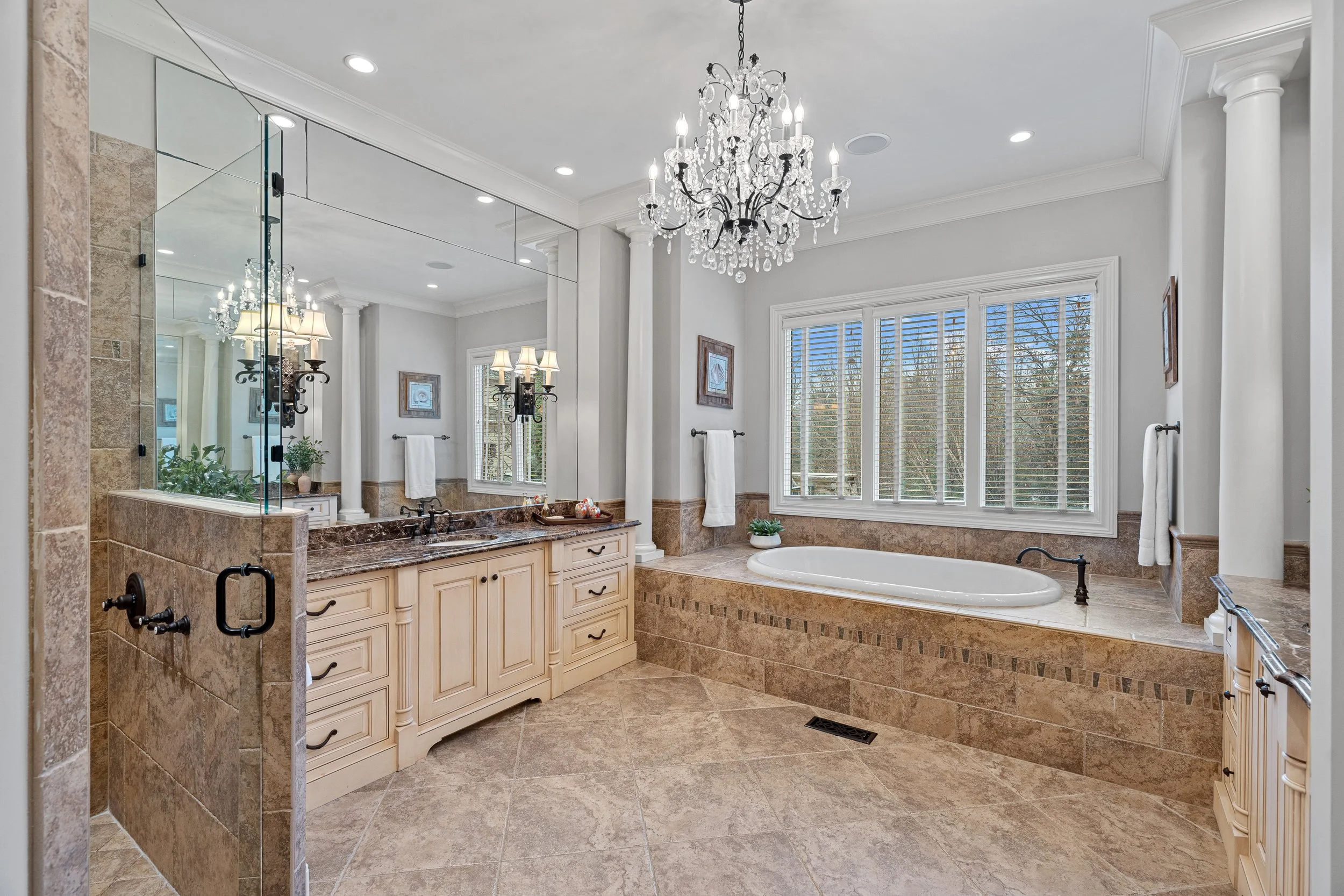 Luxury bathroom with a large bathtub by window, marble countertops, cabinetry, chandelier, and mirror.