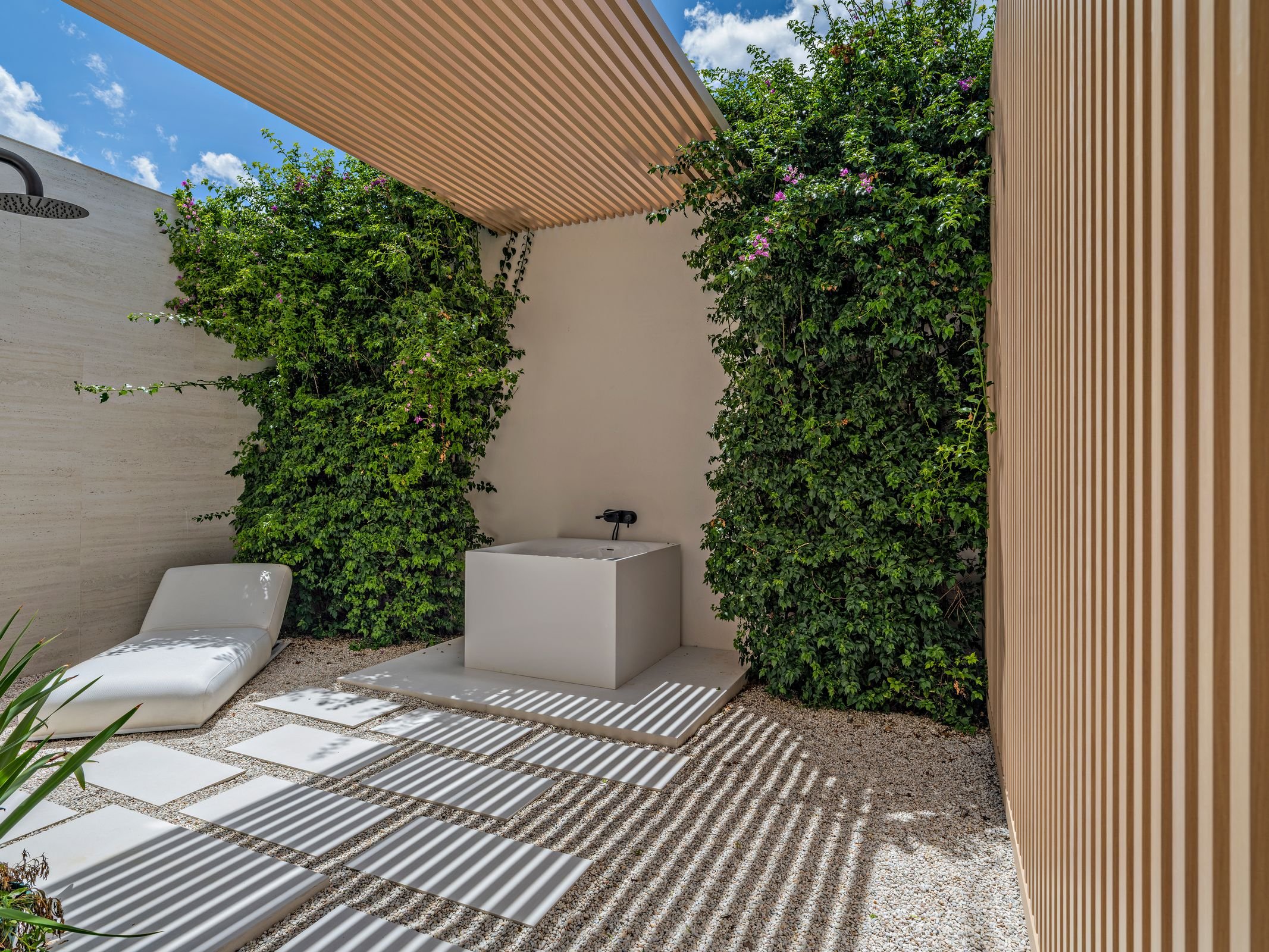 Outdoor shower area with a white lounge chair, a white sink with a black faucet, and green bushes with pink flowers. The area has a wooden slat roof casting striped shadows on the ground, which is covered with white tiles and gravel.