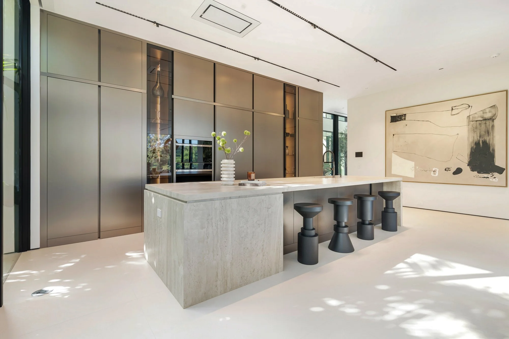 Modern kitchen with marble island, black bar stools, and sleek gray cabinetry, with abstract artwork on the wall and natural light coming through large windows.