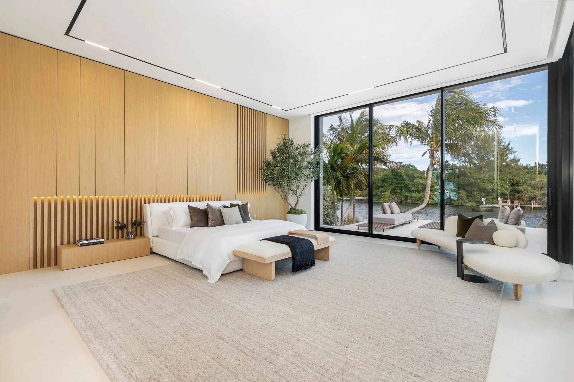 Modern bedroom with large glass windows overlooking palm trees and water, white bed with gray and brown pillows, wooden wall decor, white lounge chair, and beige rug.