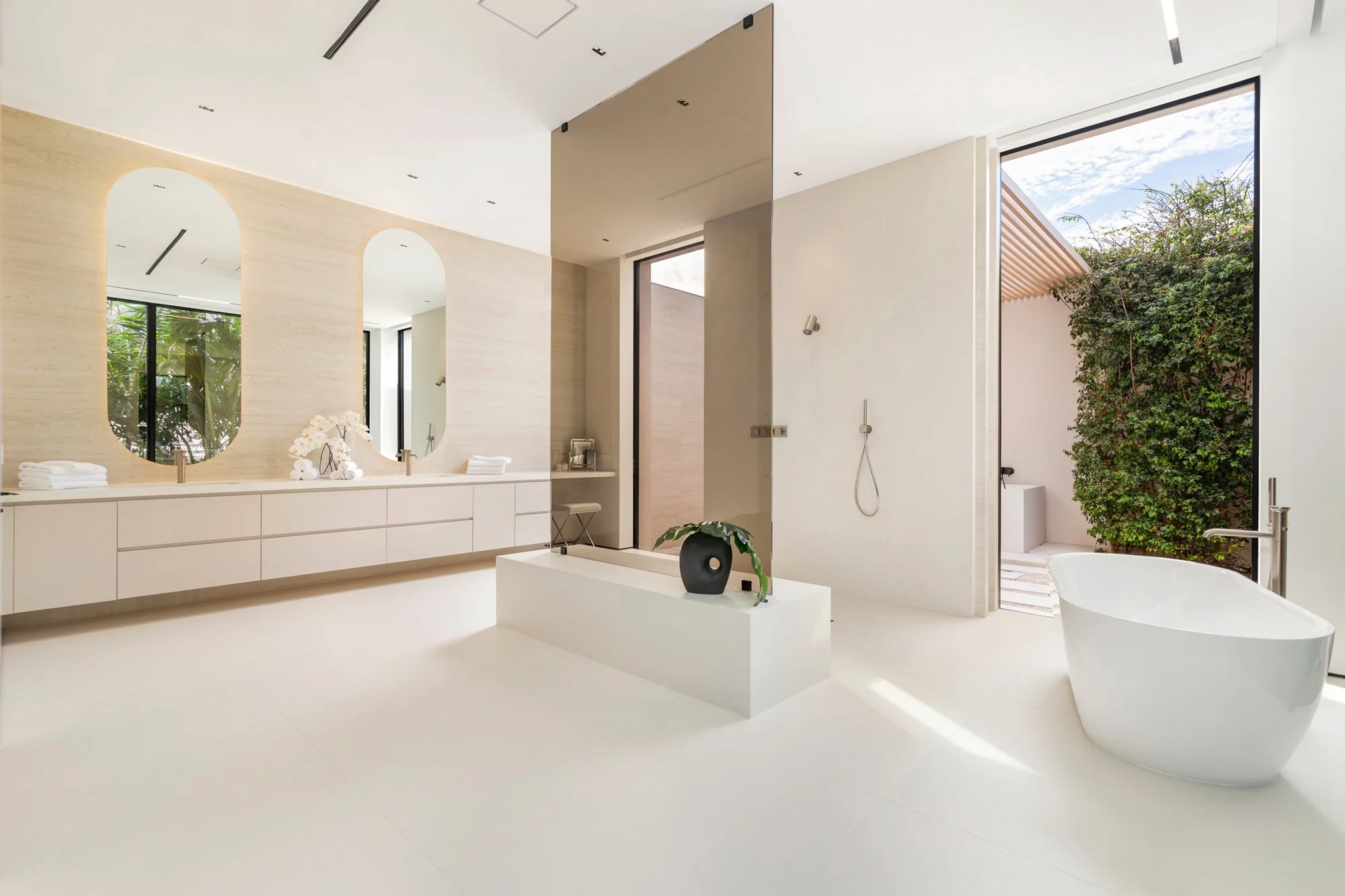 Modern, minimalistic bathroom with white walls, a freestanding bathtub, a large window with outdoor greenery, a double vanity with oval mirrors, and a neutral color palette.