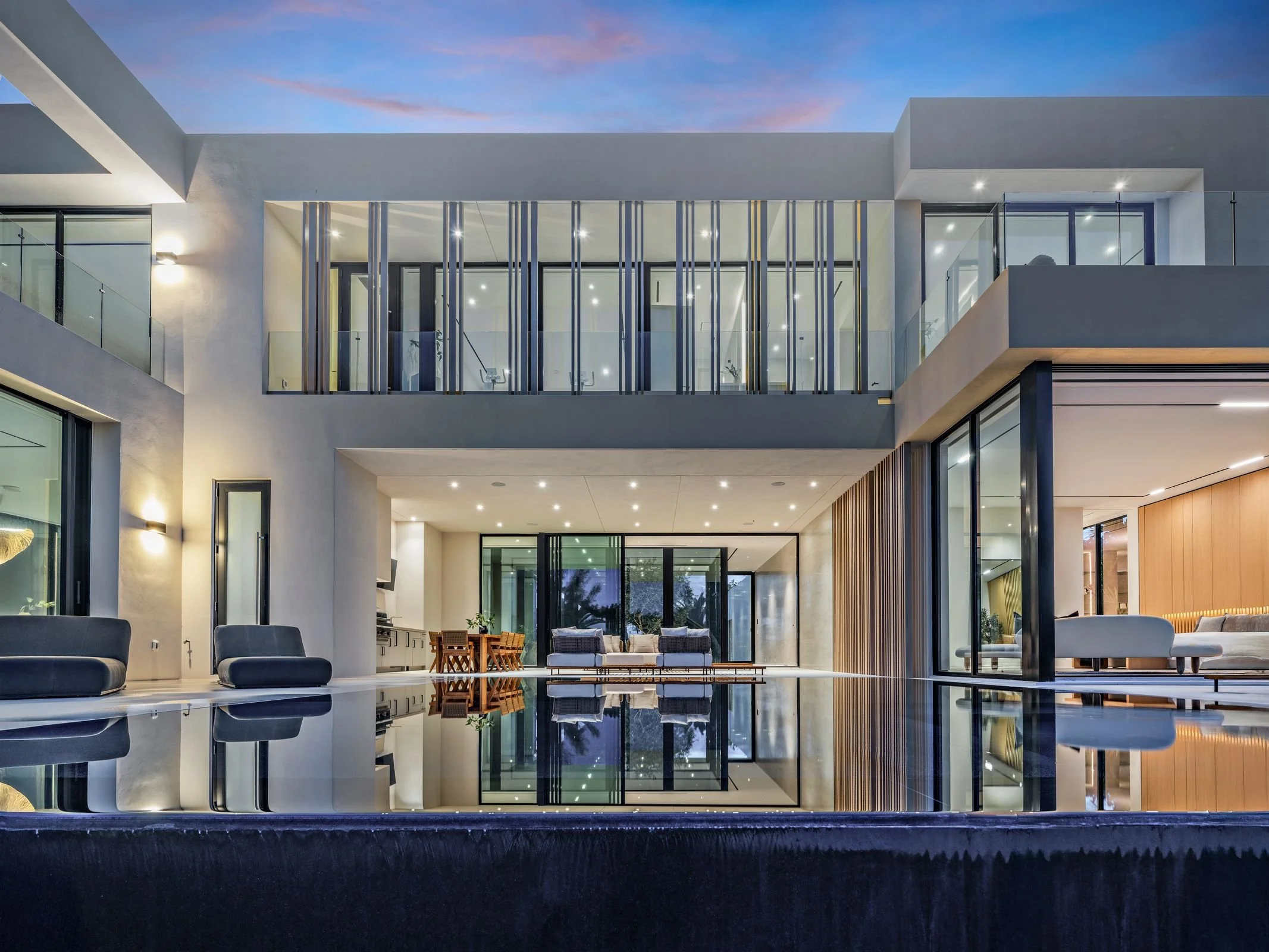 Modern multi-story house with open living area, large glass windows, and outdoor patio reflected in pool at dusk.