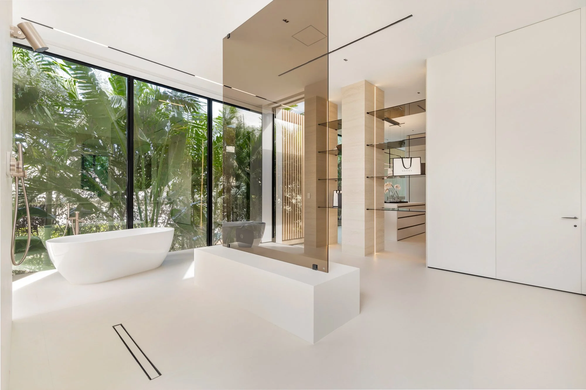 Modern bathroom with white freestanding bathtub next to large glass window overlooking lush greenery, minimalist fixtures, and neutral-toned decor.