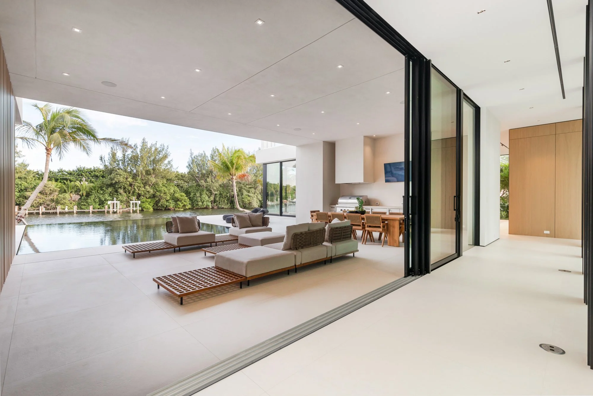 Modern living space with indoor-outdoor design, featuring a large sliding glass door, outdoor seating with lounge chairs, a dining area, and a view of a water body surrounded by lush trees and palms.