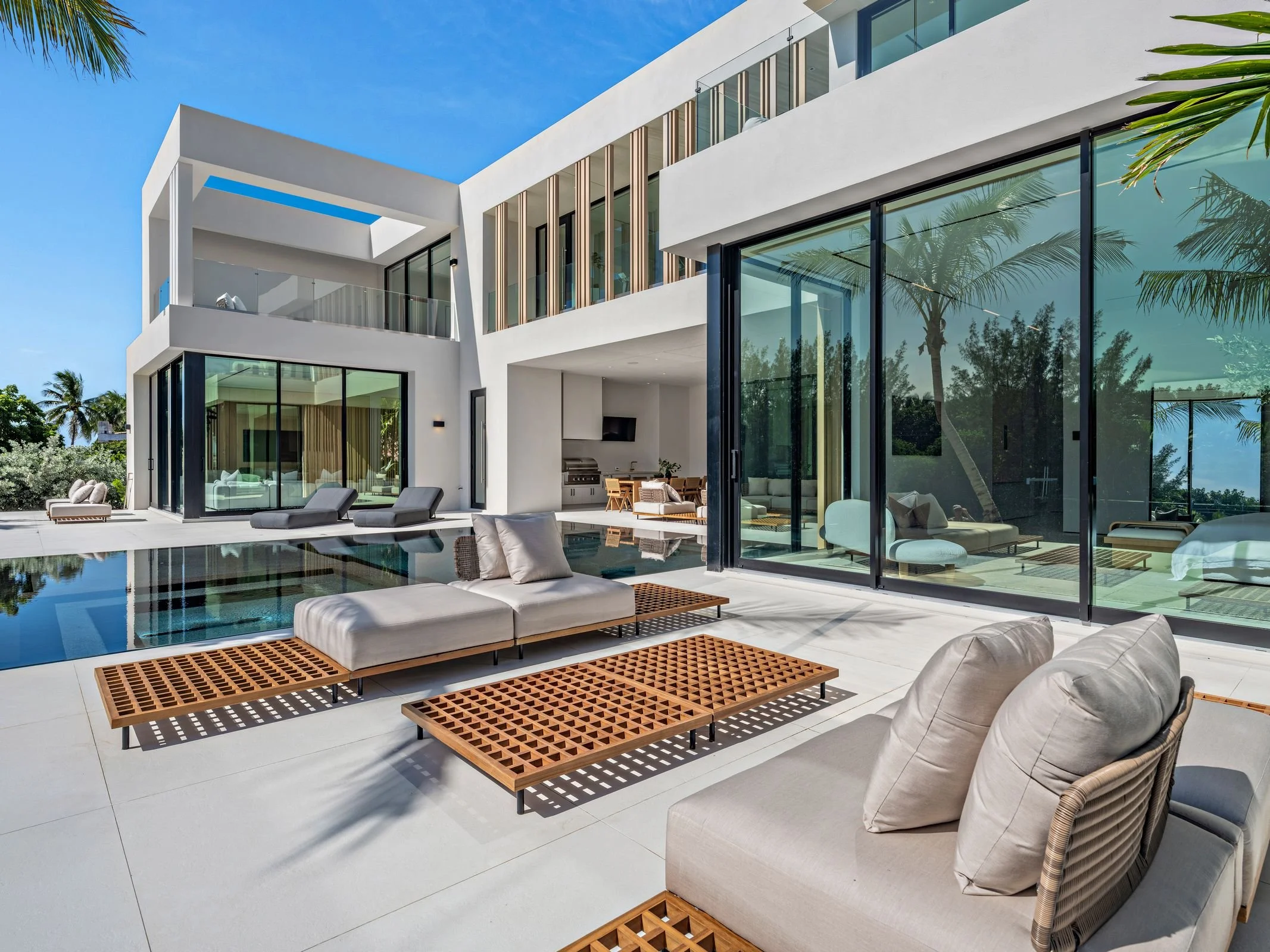 Modern white house with large glass windows and sliding doors, outdoor lounge area with cushioned chairs and wooden tables, swimming pool, palm trees, under a clear blue sky.
