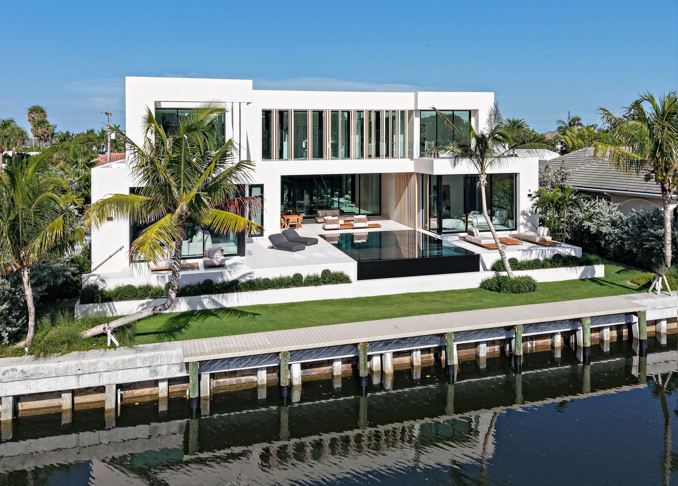 Modern white house with large glass windows, surrounded by palm trees, overlooking water with dock, featuring outdoor seating and a swimming pool.