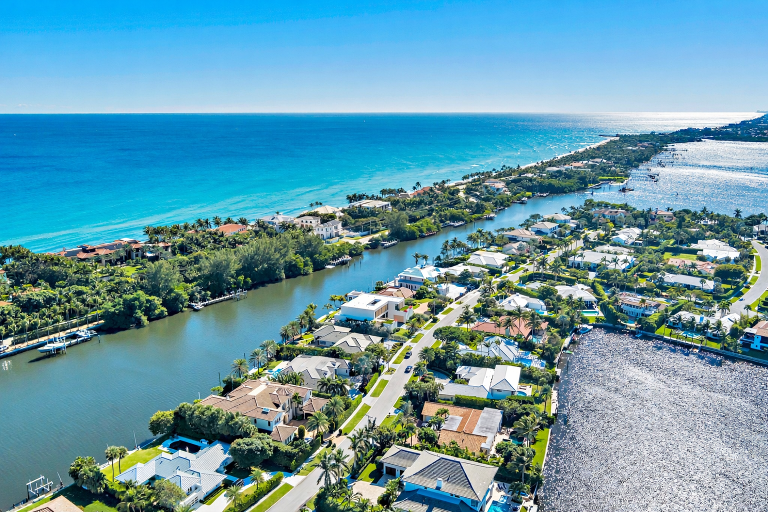 Aerial view of a residential neighborhood near the coast with houses, water canals, and the ocean in the background under a clear blue sky.
