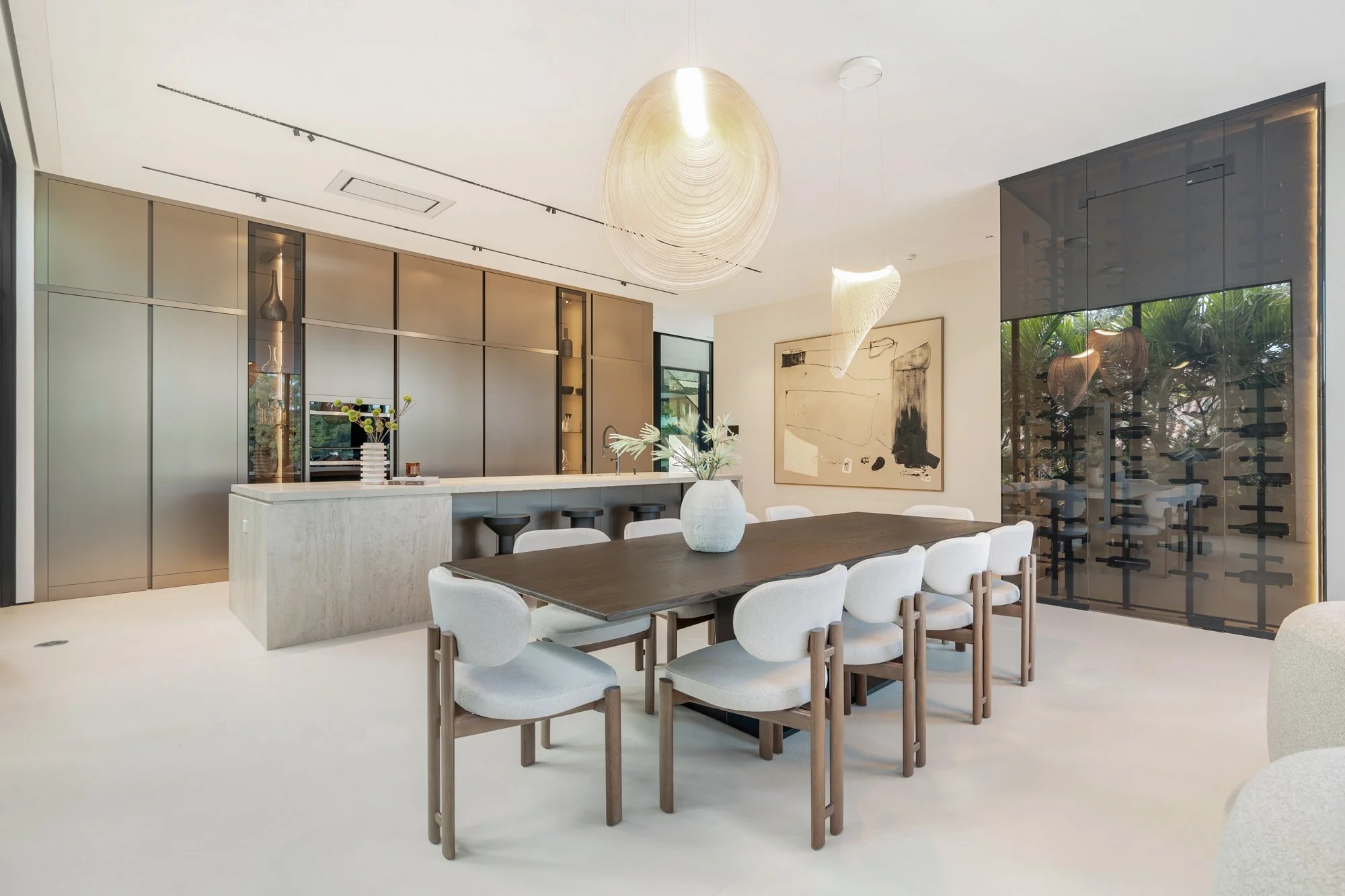 Modern dining room with a large wooden table, white upholstered chairs, a vase with flowers, and a minimalist art piece on the wall. A kitchen bar with stools and sleek cabinetry in the background. Elegant lighting fixtures hang from the ceiling, and