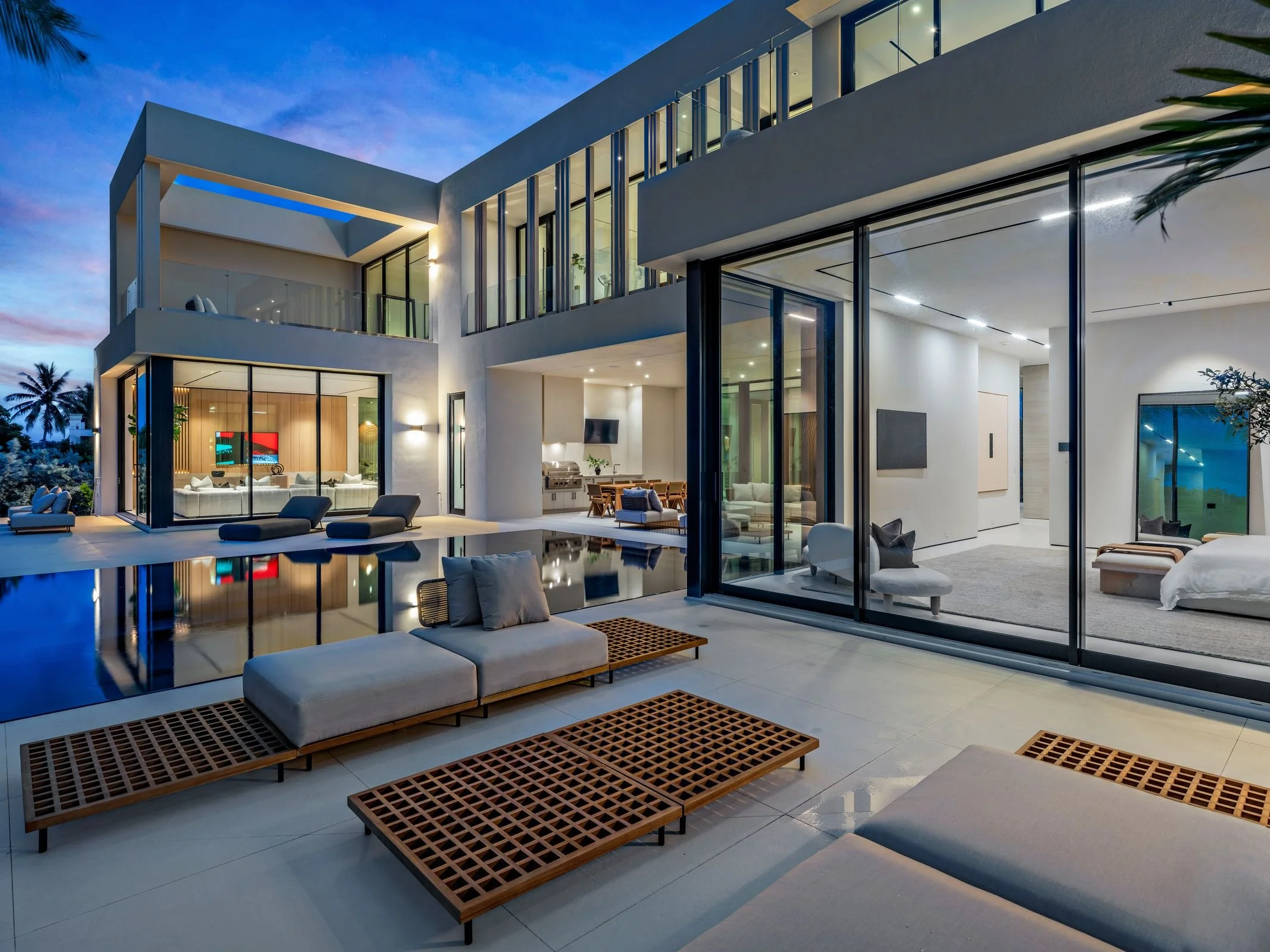 Modern house with a swimming pool and outdoor lounge area at dusk, with large glass windows revealing the interior living space.