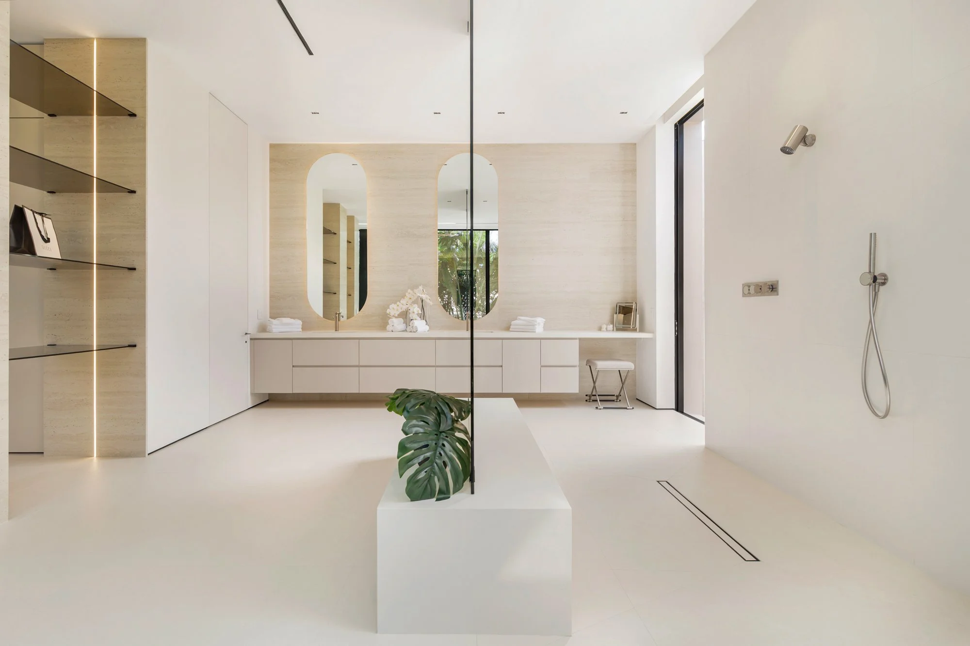 Minimalist bathroom with a large vanity mirror, white walls, and a shower area on the right with a handheld showerhead. There are towels, decor, and a chair on the vanity counter.