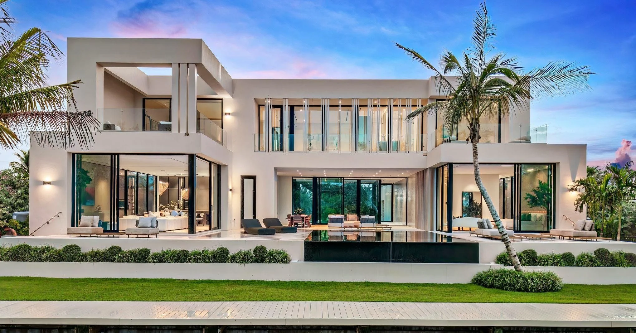 Modern white multi-story house with large glass windows, palm trees, and a landscaped yard with green grass and bushes.