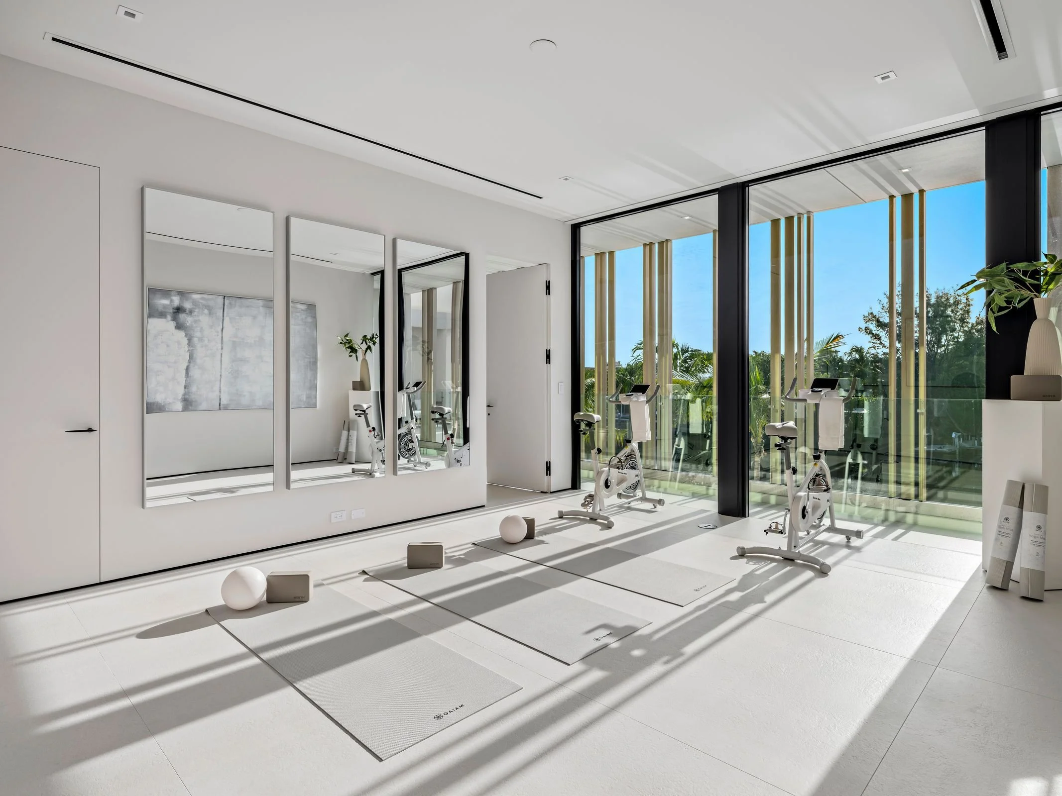 Modern home gym with exercise bikes, yoga mats, and sunlight streaming through large floor-to-ceiling windows with outdoor greenery.