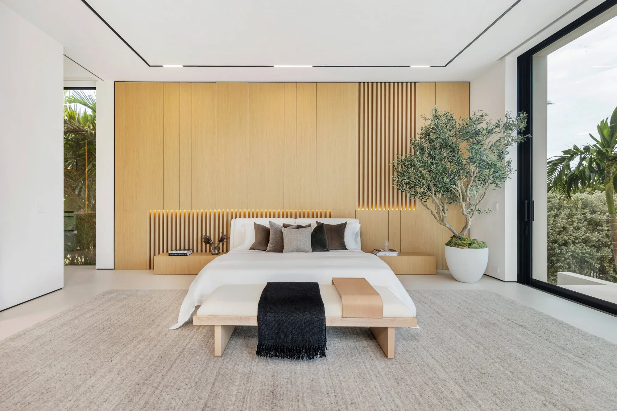 Modern bedroom with a wooden accent wall, large window with outside greenery, white bed with gray and brown pillows, beige bench with black and beige throws, large potted tree, and minimalist décor.