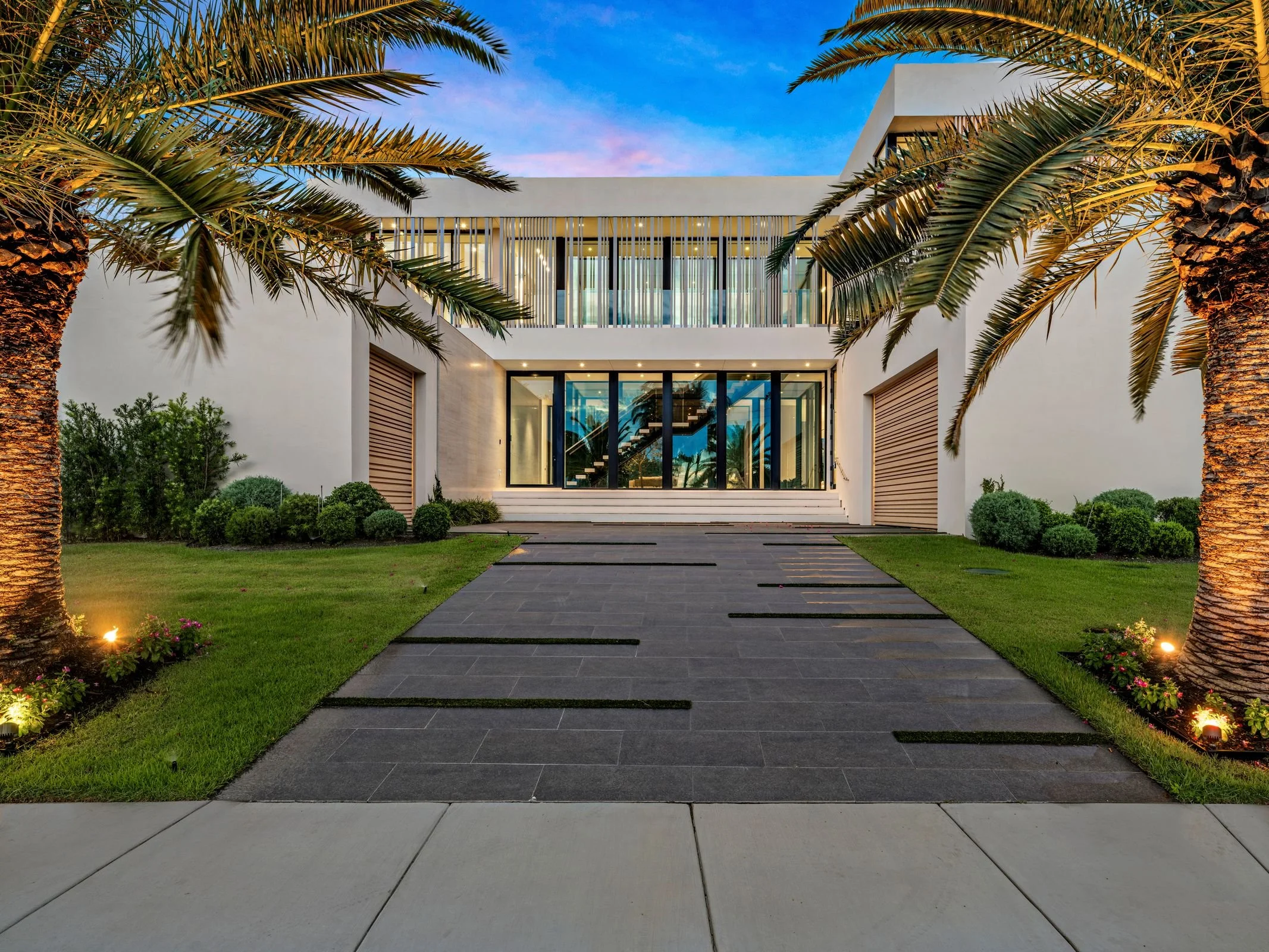 Modern white house with large glass windows, leading to a staircase inside, surrounded by a well-maintained garden and palm trees at dusk.