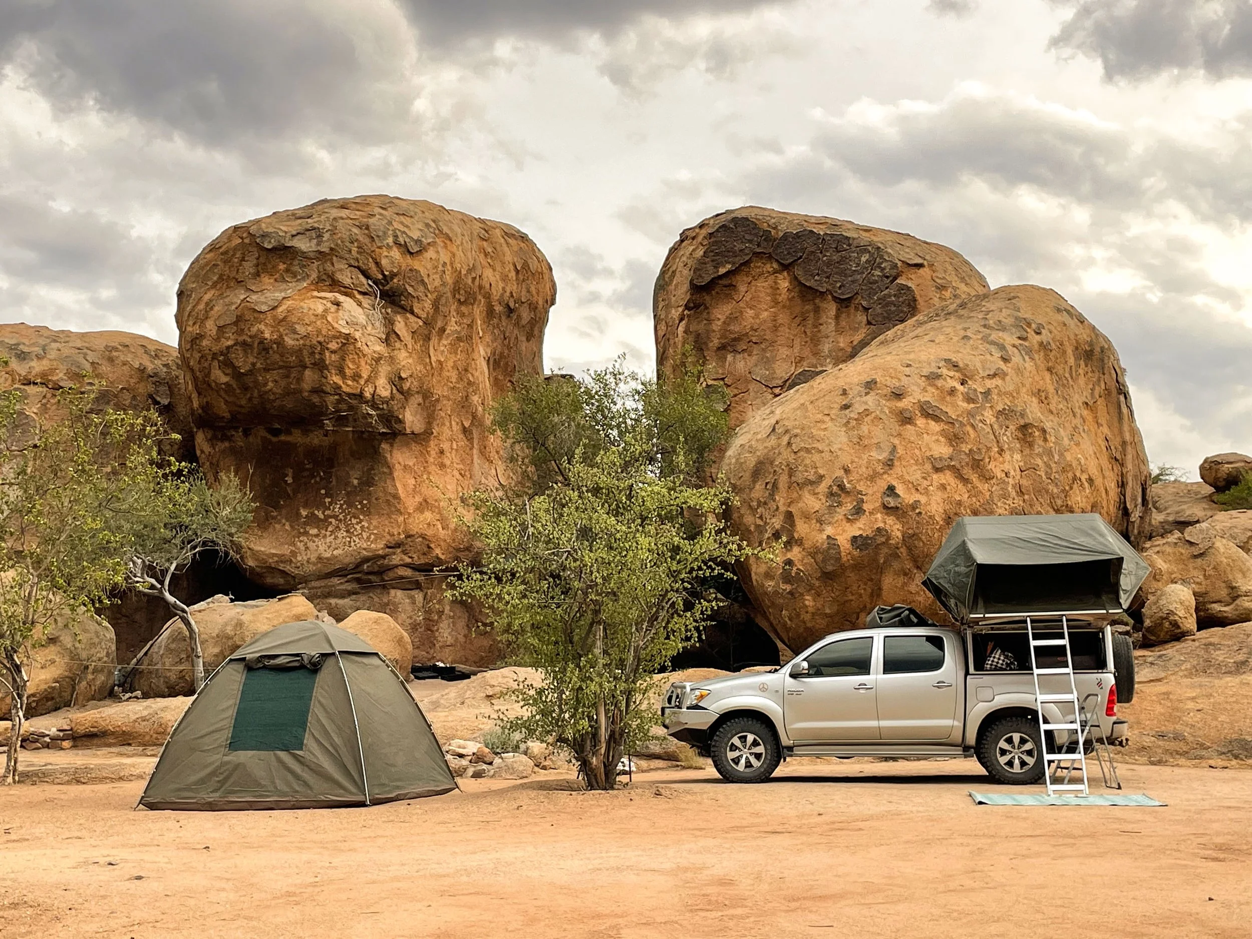 Campingplatz mit einem Zelt und einem Pickup-Truck in der Wüstenlandschaft, umgeben von großen Felsen und Bäumen. Rooftop tent trip Namibia