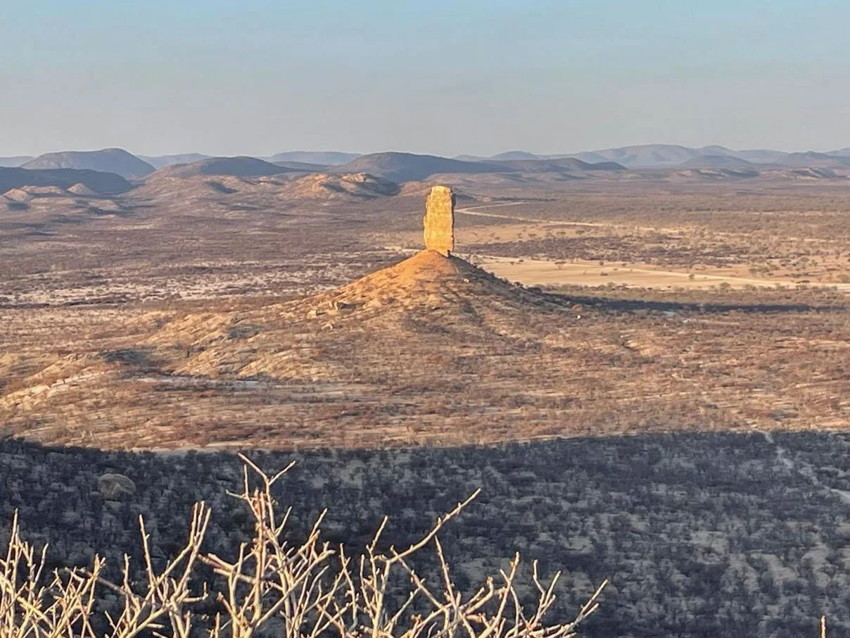 Ein einzelner, hoher, fast rechteckiger Steinkoloss auf einem Hügel in einer Wüstenlandschaft mit Bergen im Hintergrund. Vingerklip namibia